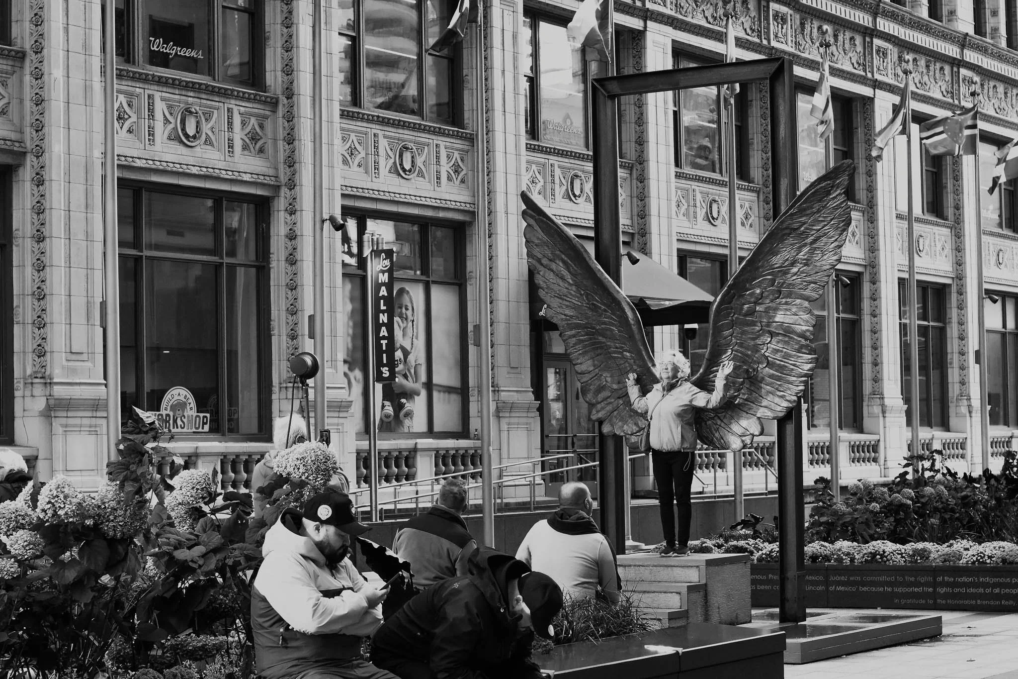 A woman with butterfly wings standing on a small stage, speaking or singing in front of an audience outside a historic building.