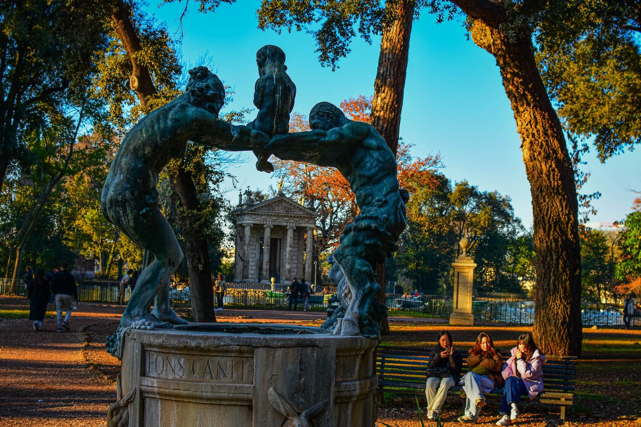 The photo shows a park with a bronze sculpture of two boys playing, one holding a smaller child up in the air. The sculpture is on a stone pedestal. In the background, there are trees with green and orange leaves, suggesting fall. Several people are 