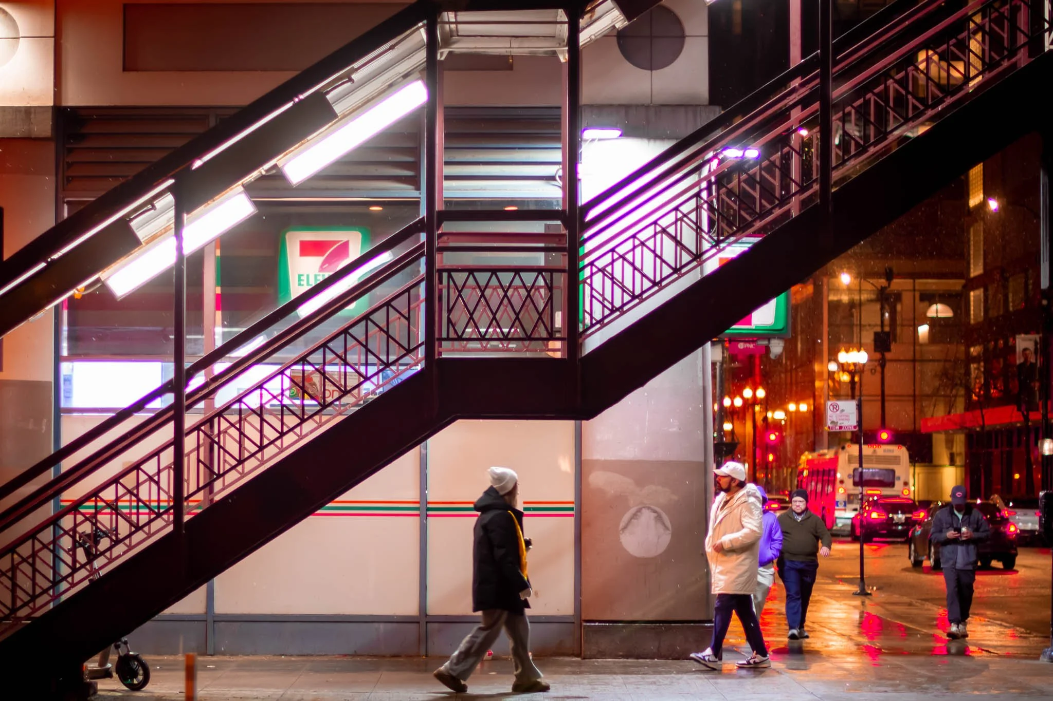 Nighttime street scene with pedestrians walking past a 7-Eleven store, illuminated signs, and traffic. Metal staircase with neon lights is in the foreground.