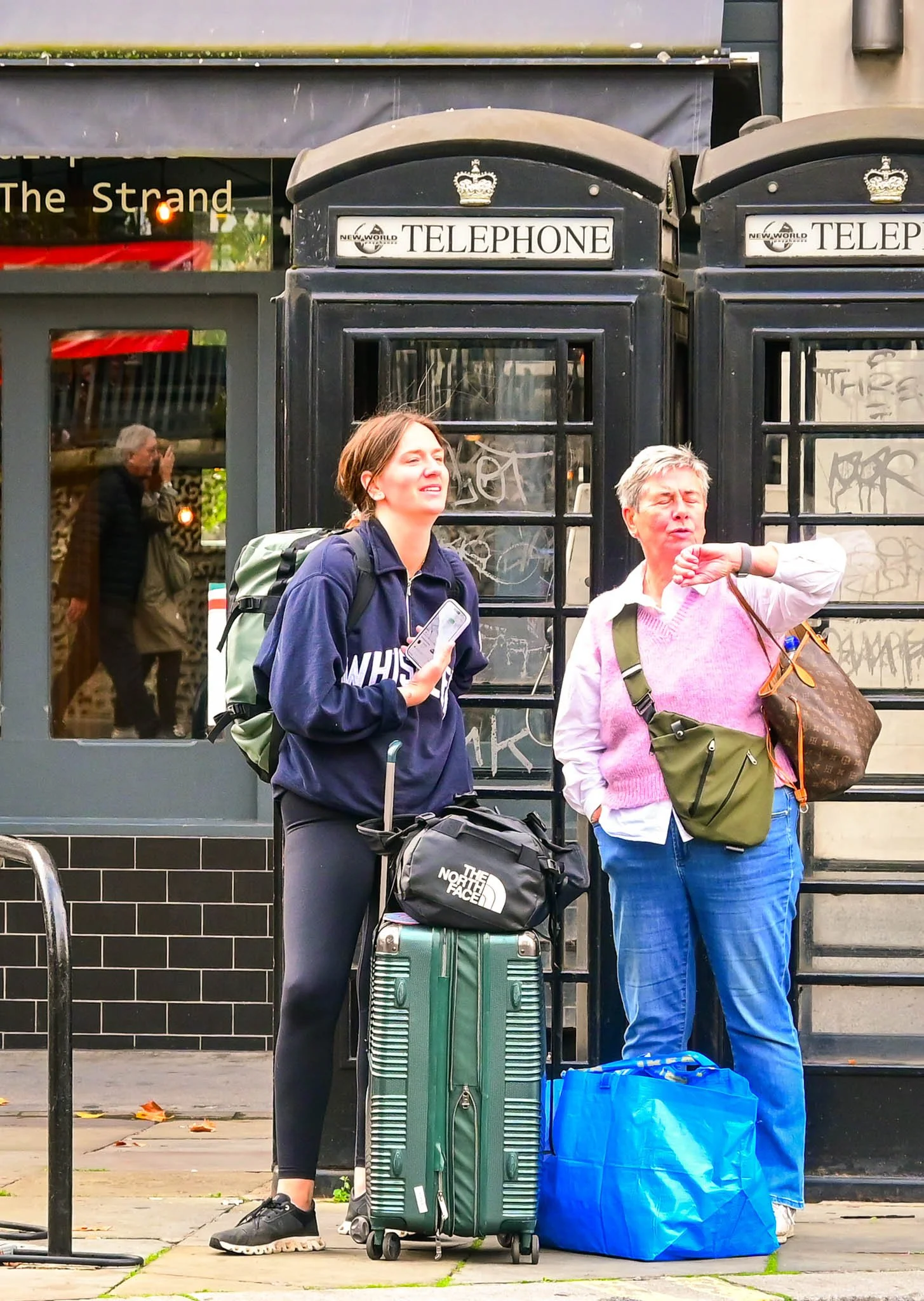 Two women with luggage standing in front of traditional British telephone booths, possibly waiting or talking.