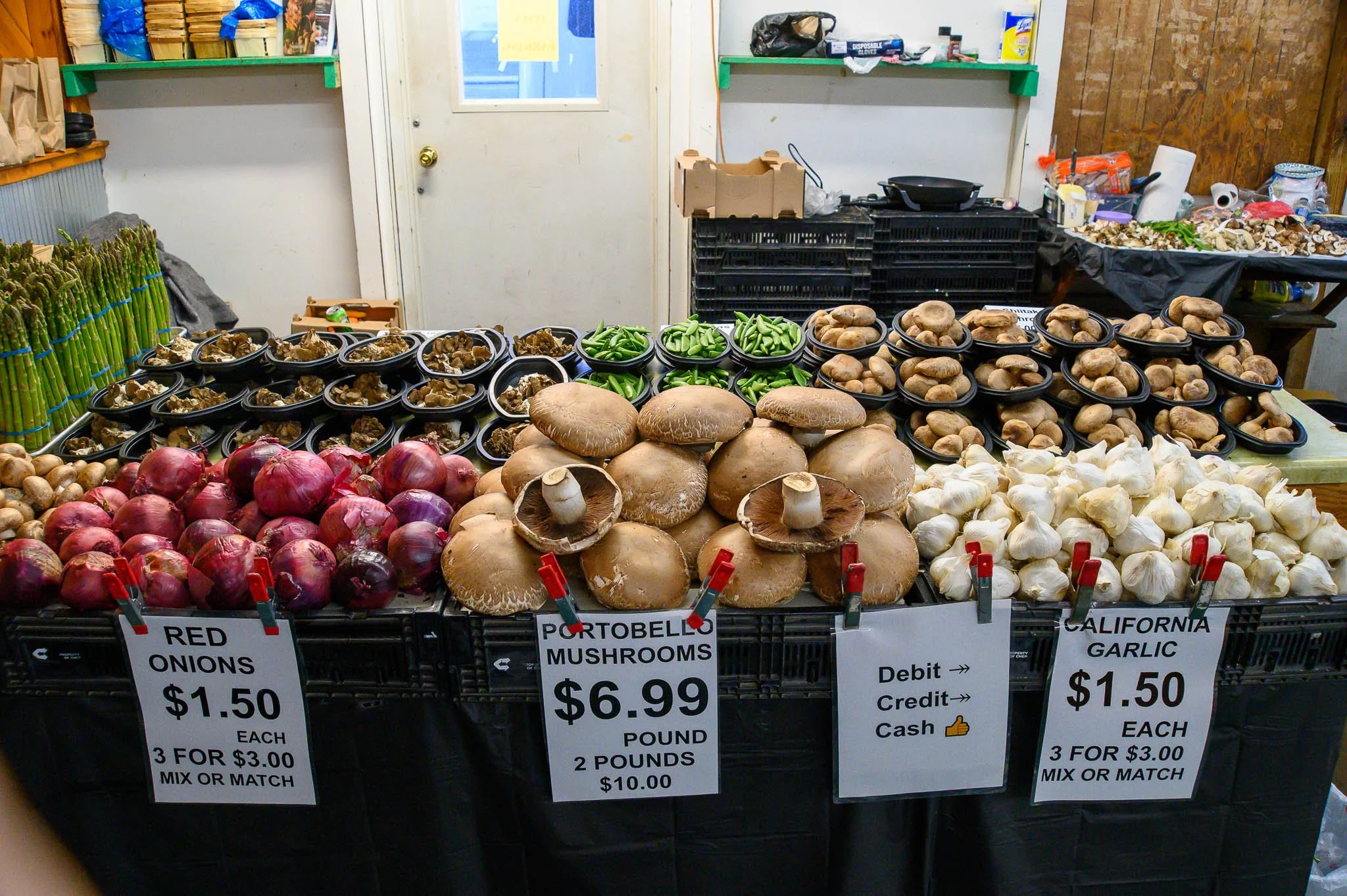Fresh produce at a farmers market stand, including red onions, portobello mushrooms, garlic, and green beans, with price signs and a sign indicating payment options.