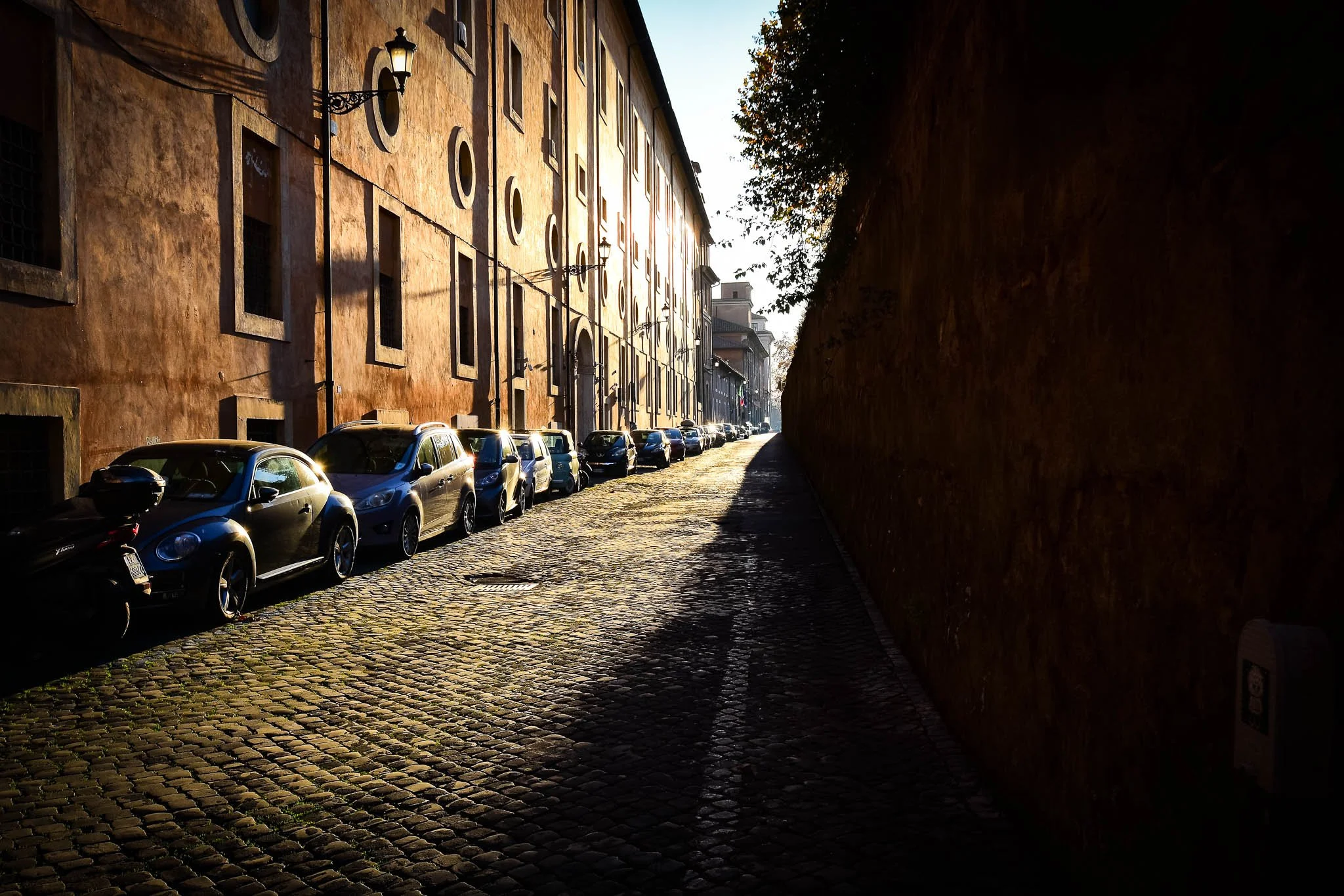 A cobblestone street with parked cars along a building wall, with sunlight casting shadows and illuminating the scene.