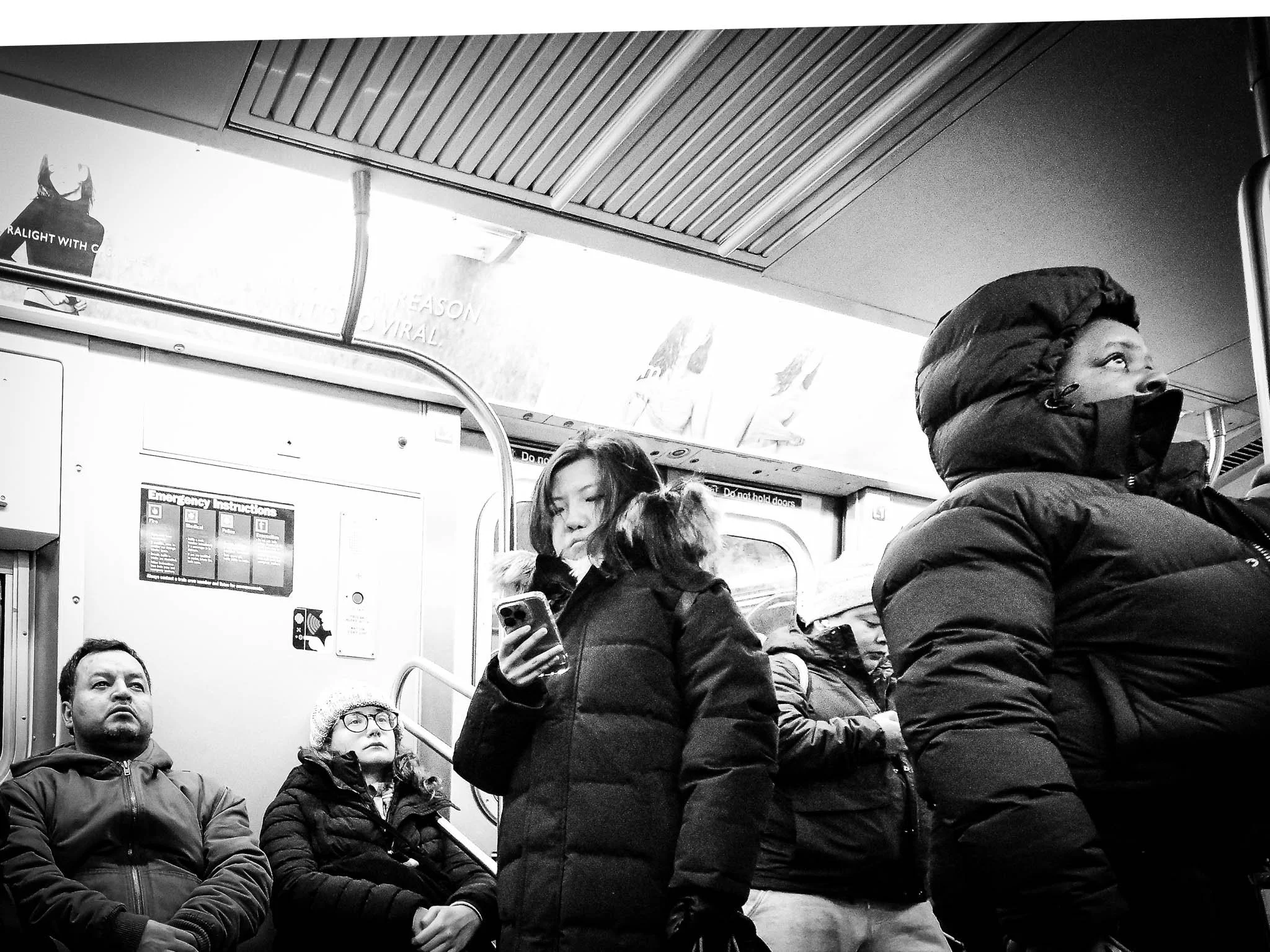 People standing and sitting on a subway train, bundled in winter clothing, with some looking at their phones and others looking forward or down.