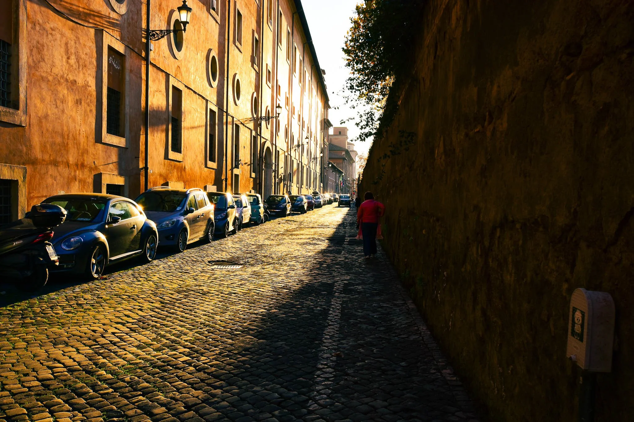 Cobblestone street with parked cars on the left and a stone wall on the right, with a pedestrian walking in the distance during sunset.