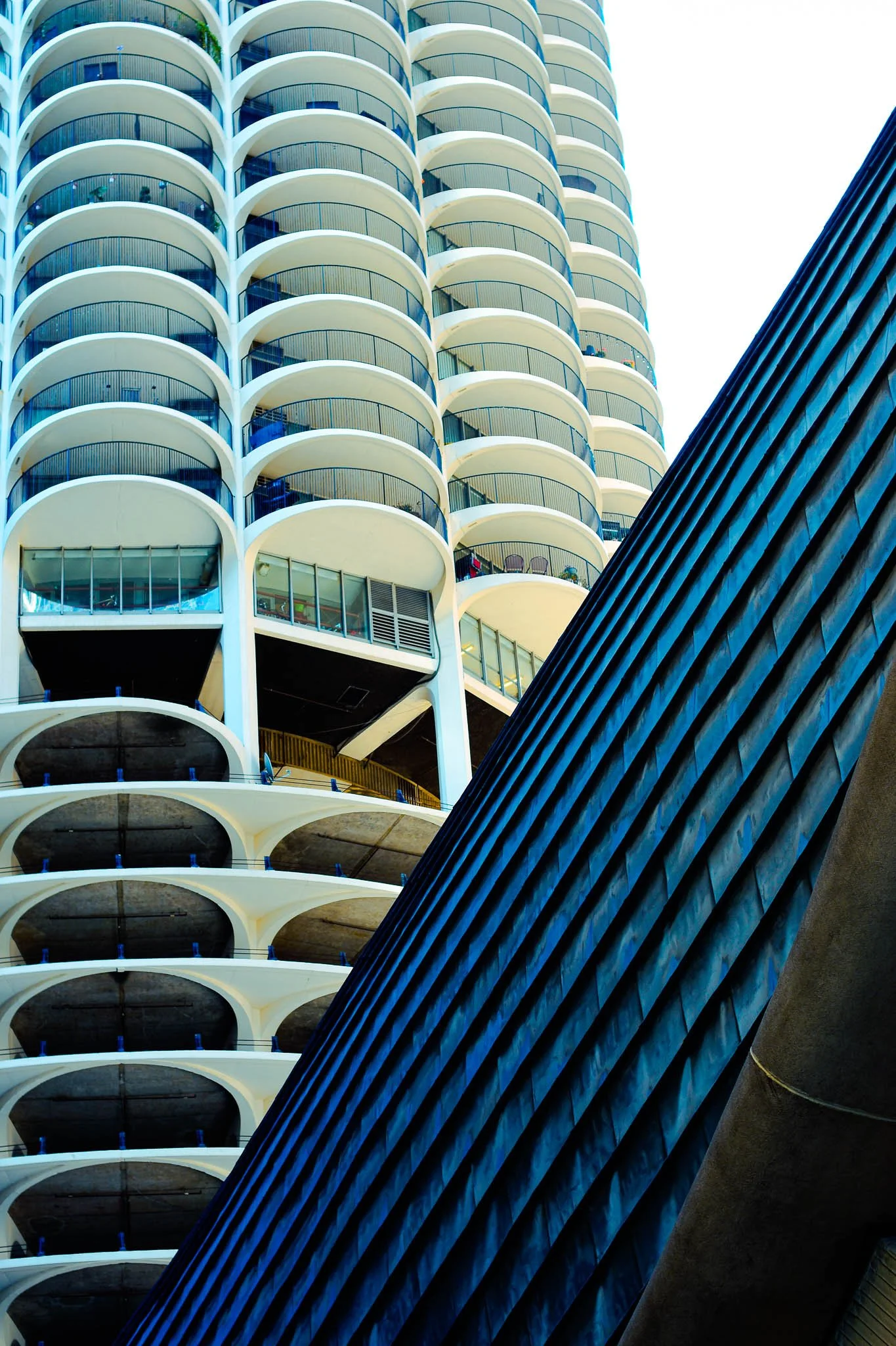 Close-up view of a modern high-rise building with rounded balconies, and part of another building with a ridged, dark blue, textured facade.