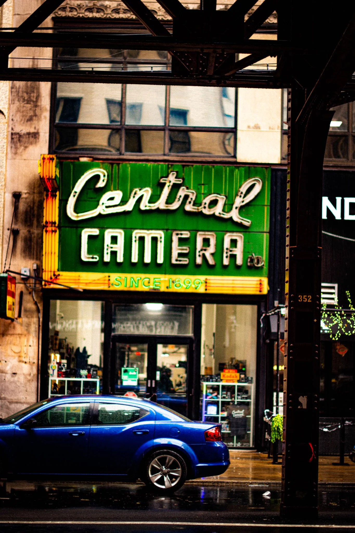 Neon sign for Central Camera, established in 1899, on the exterior of a camera store, with a blue car parked in front, under an elevated structure on a city street.