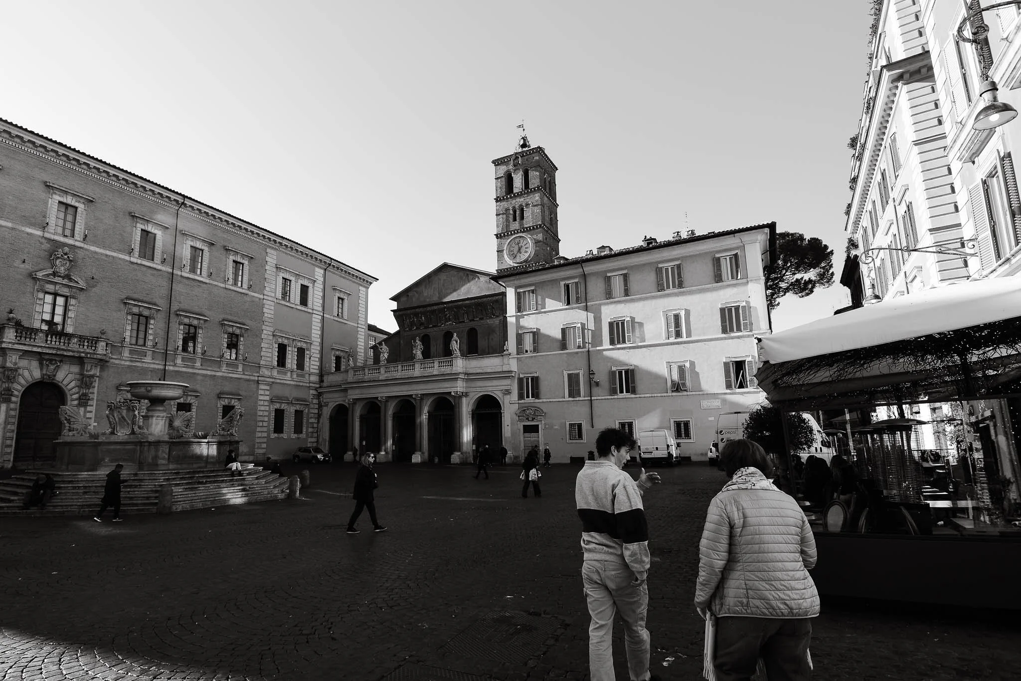 A black and white image of a historic square with a clock tower, ornate fountain, and old buildings; people are walking and talking in the square.