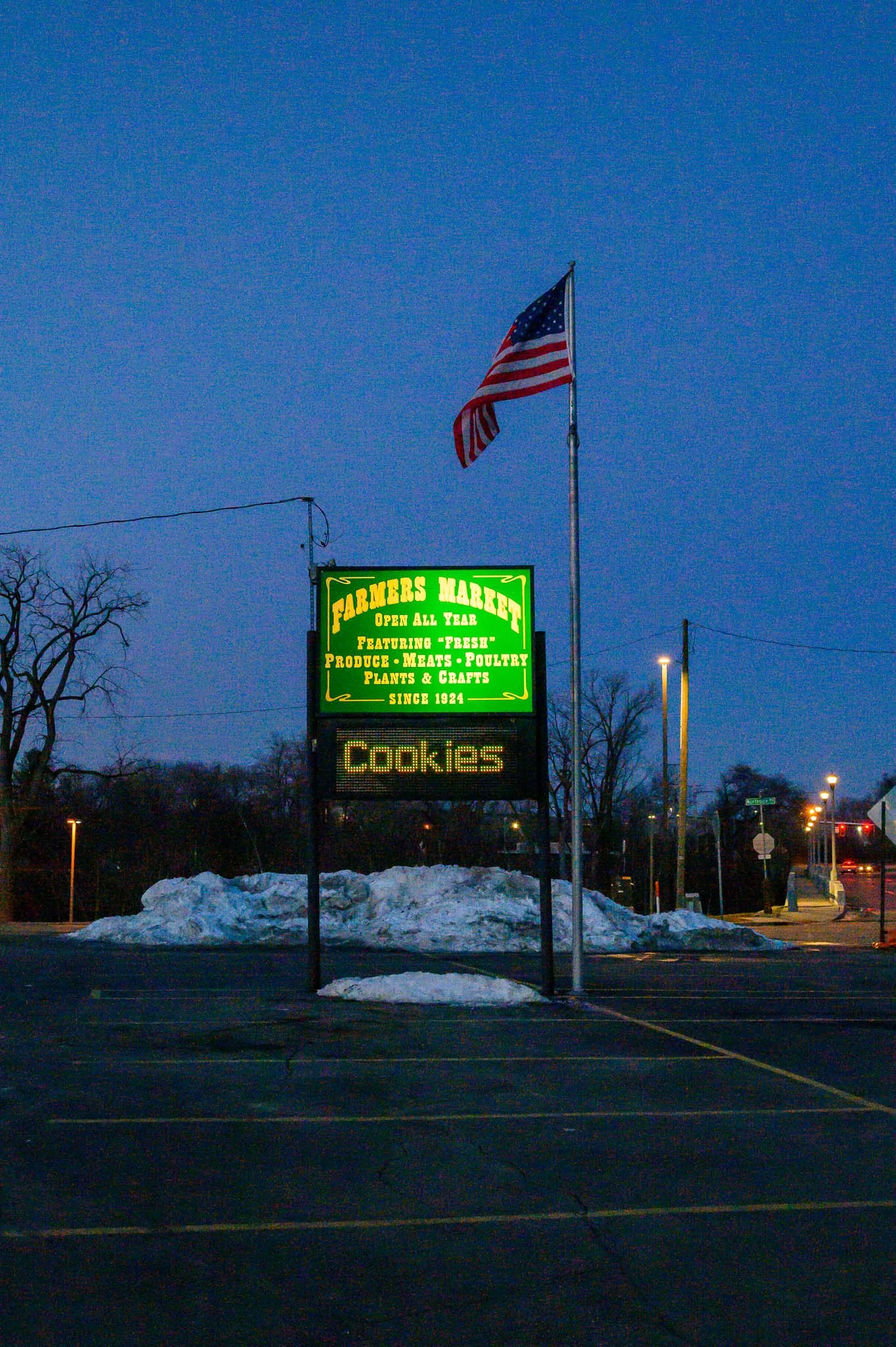 A sign outside Farmers Market, featuring produce, meats, poultry, plants, and crafts, with an electronic display reading 'Cookies'. An American flag is flying on a flagpole next to the sign, with snow on the ground in a parking lot during dusk.