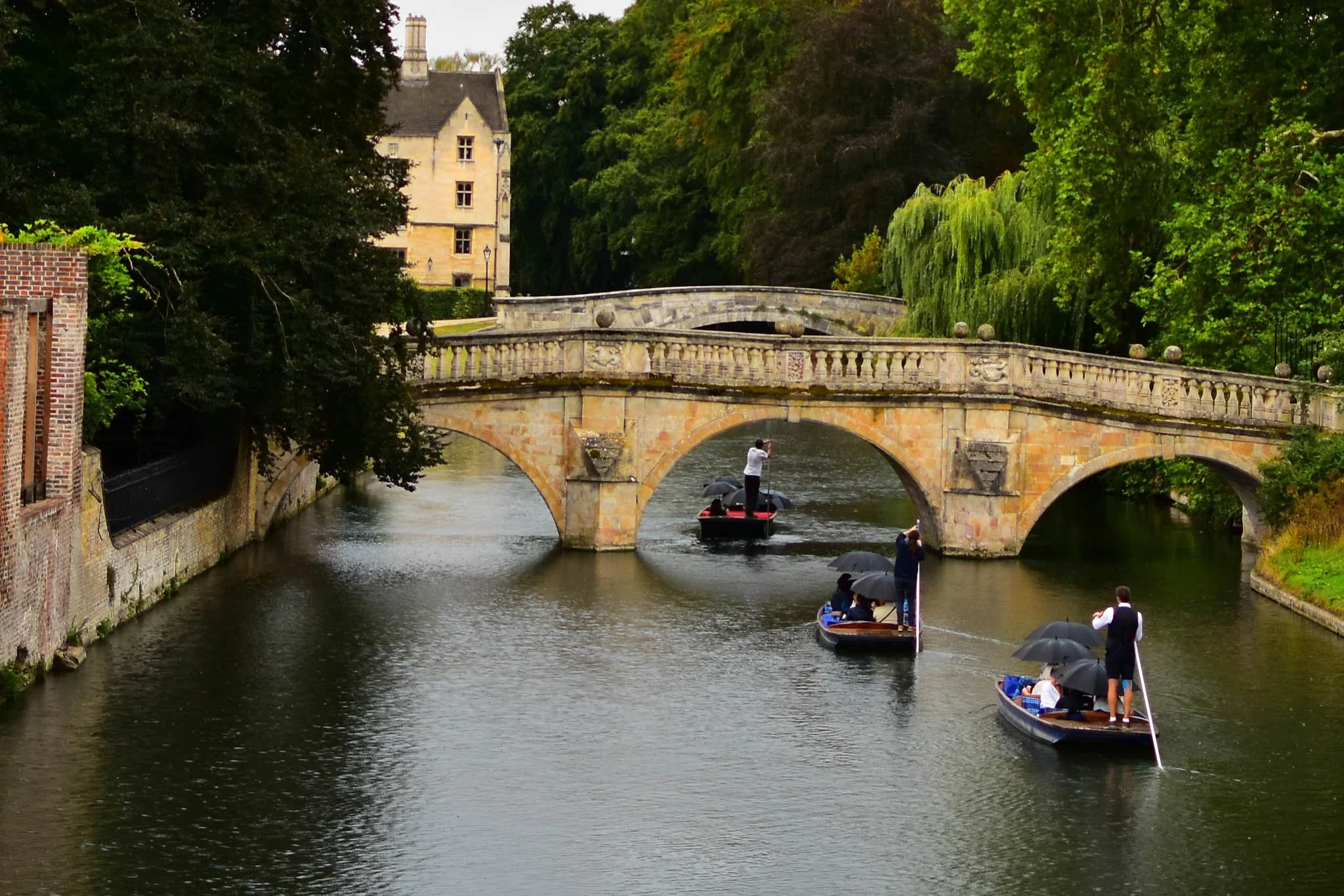 Three boats with passengers and umbrellas on a river passing under a stone bridge with decorative balustrade, surrounded by green trees and a historic building in the background.