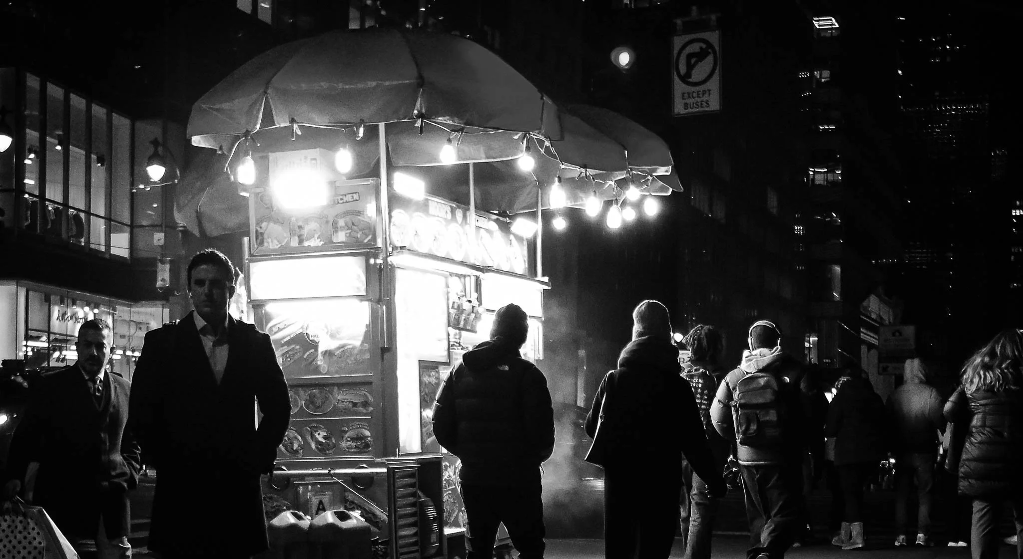 Night scene of a street food cart with bright lights, surrounded by people walking.