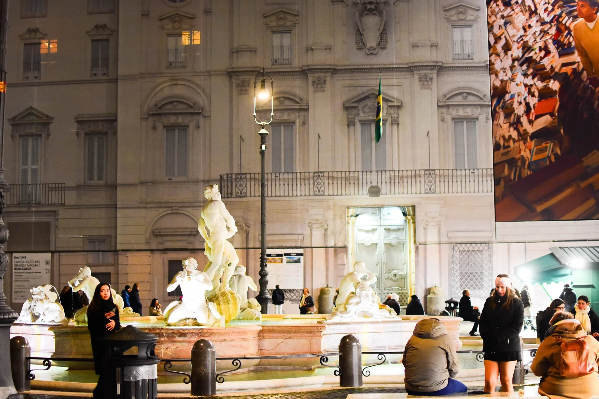 People gathered around a fountain with sculpture in front of a historic building at night. The building has a flag, windows, and a large digital billboard on the side.