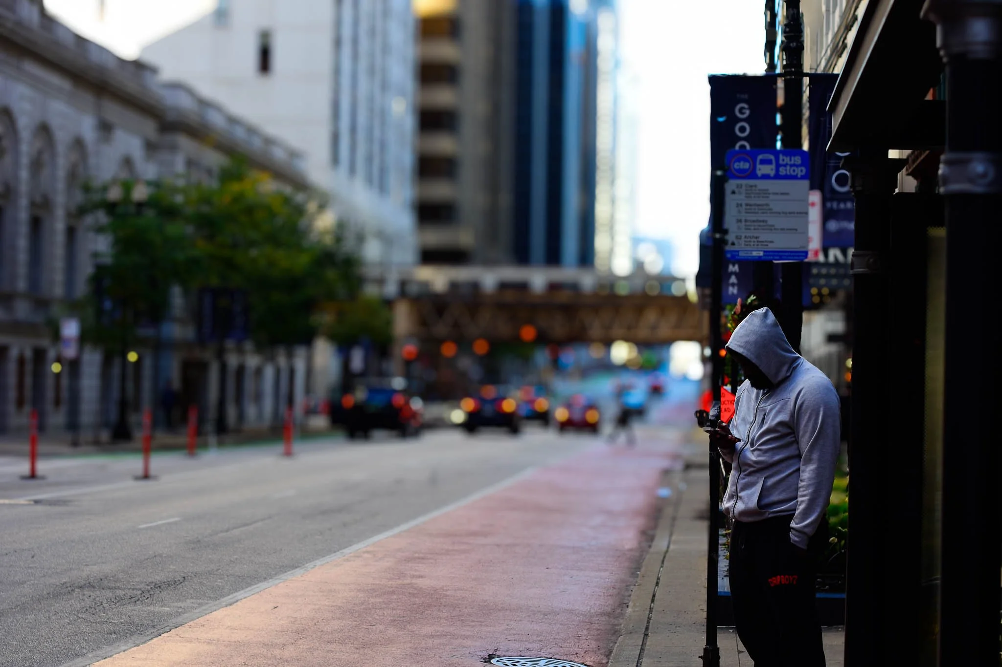 A person in a gray hoodie standing at a bus stop on a city sidewalk, looking at their phone with taller buildings in the background.