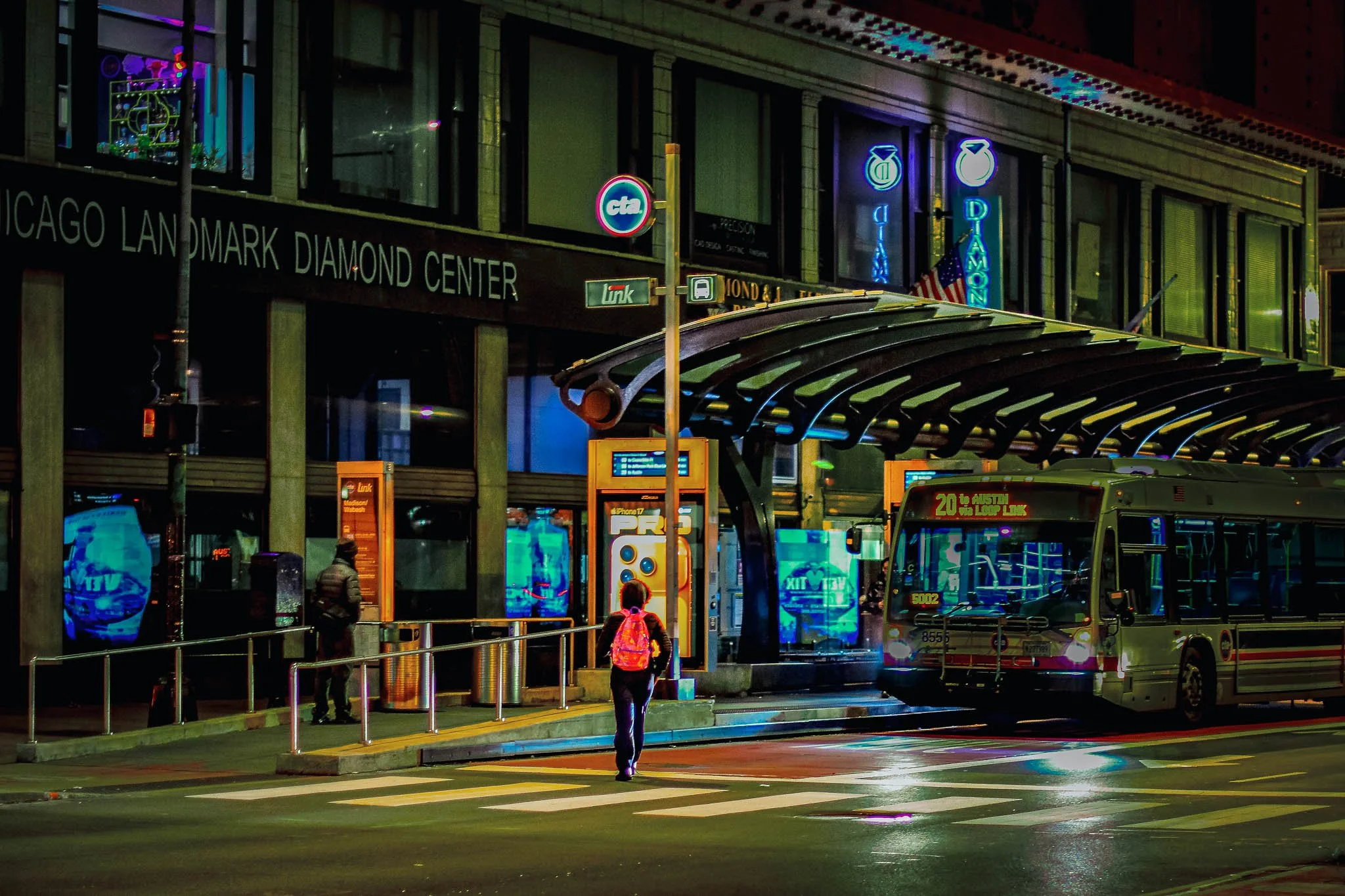 Nighttime scene outside Chicago Landmark Diamond Center with a bus parked at the bus stop, two pedestrians walking, illuminated signs, and reflections on the wet street.