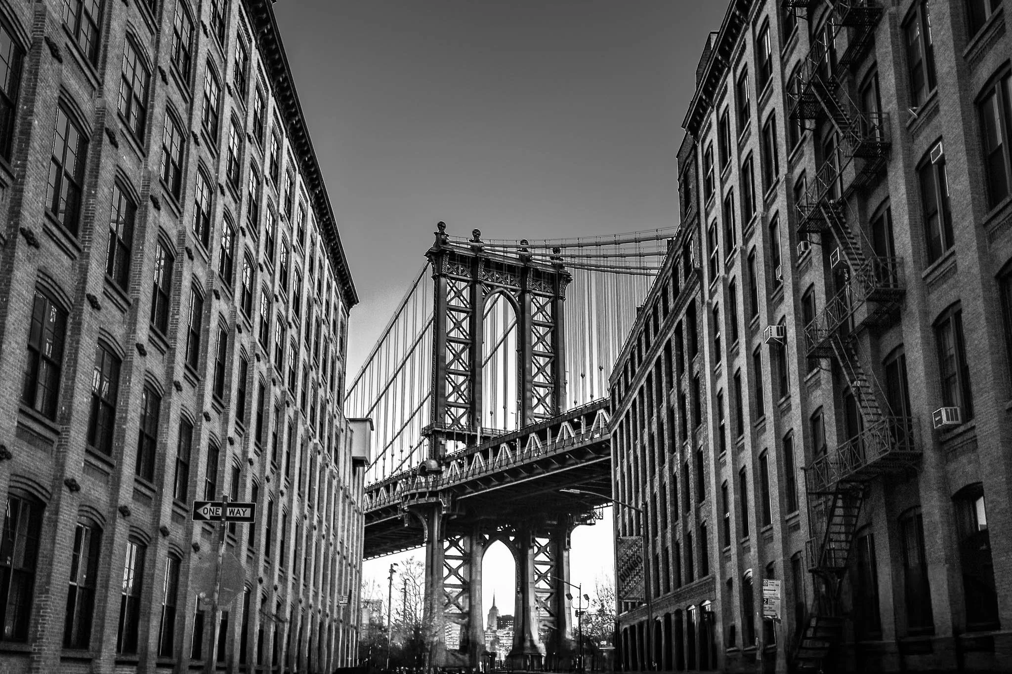 Black and white photo of Manhattan Bridge viewed from between two brick buildings in Brooklyn, New York City.