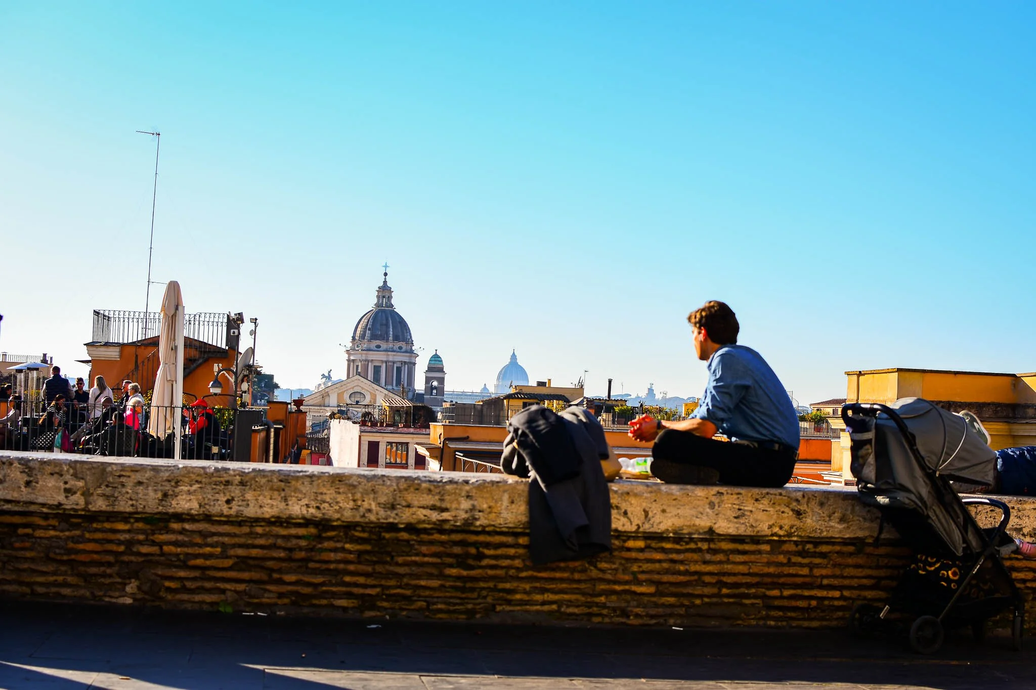 A man sitting on a rooftop ledge overlooking Rome, with church domes in the distance, during daytime.