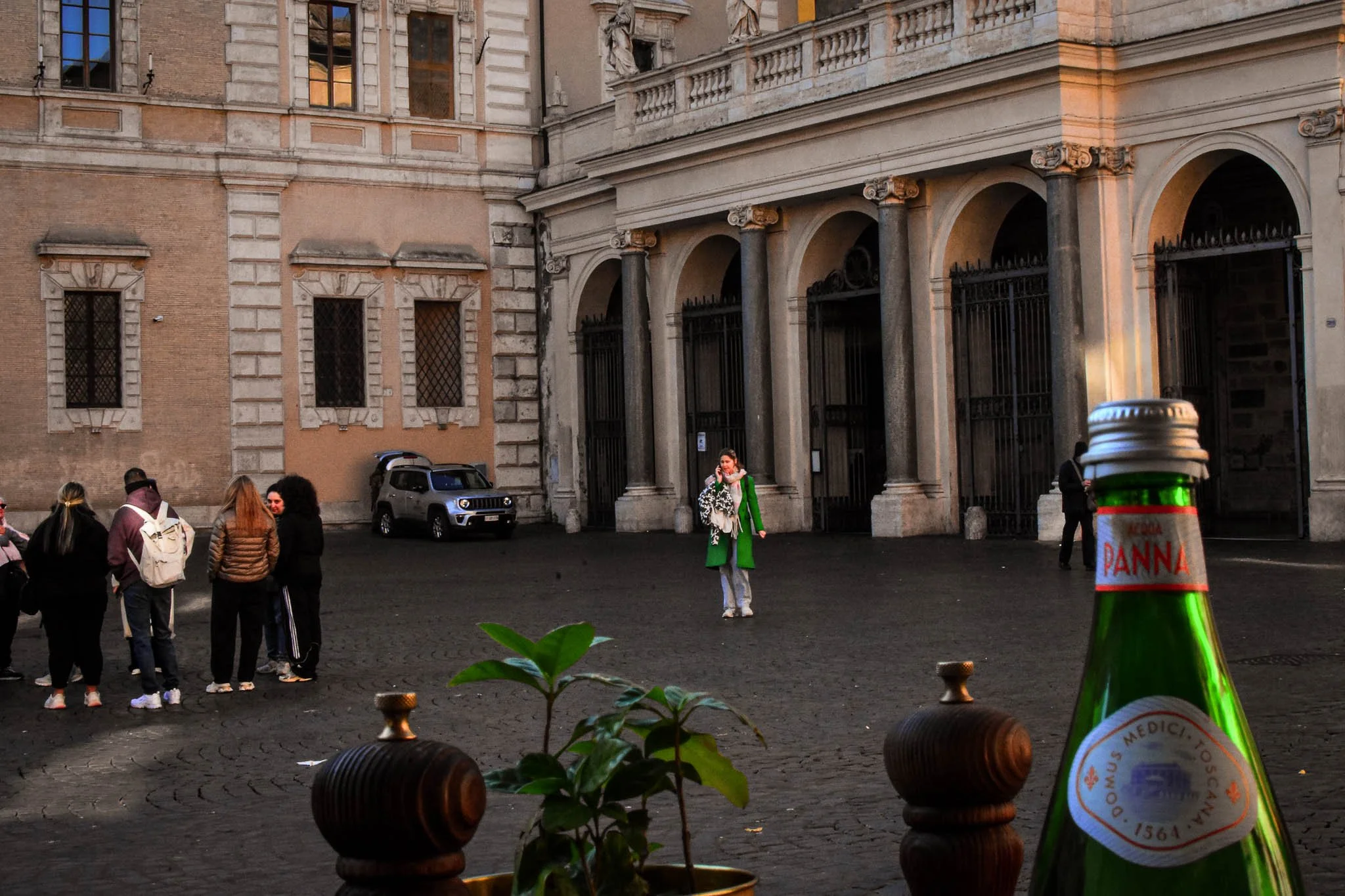 A woman in a green coat standing in a cobblestone courtyard among a group of people, with historic buildings in the background and a large green bottle of Panna mineral water in the foreground.