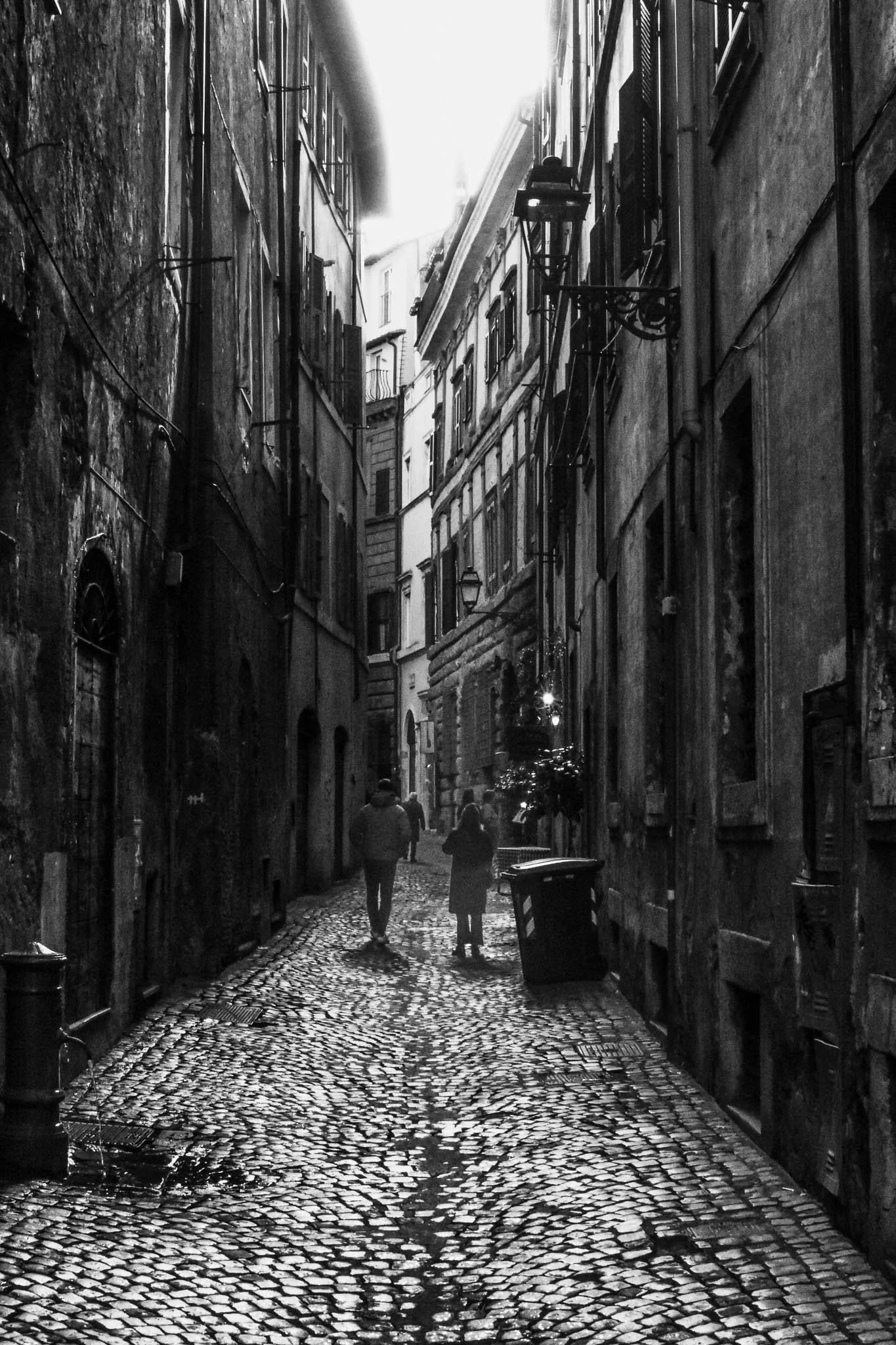 A narrow black and white alleyway with cobblestone pavement, flanked by old weathered buildings with multiple windows and fire escapes, and three people walking down the street.