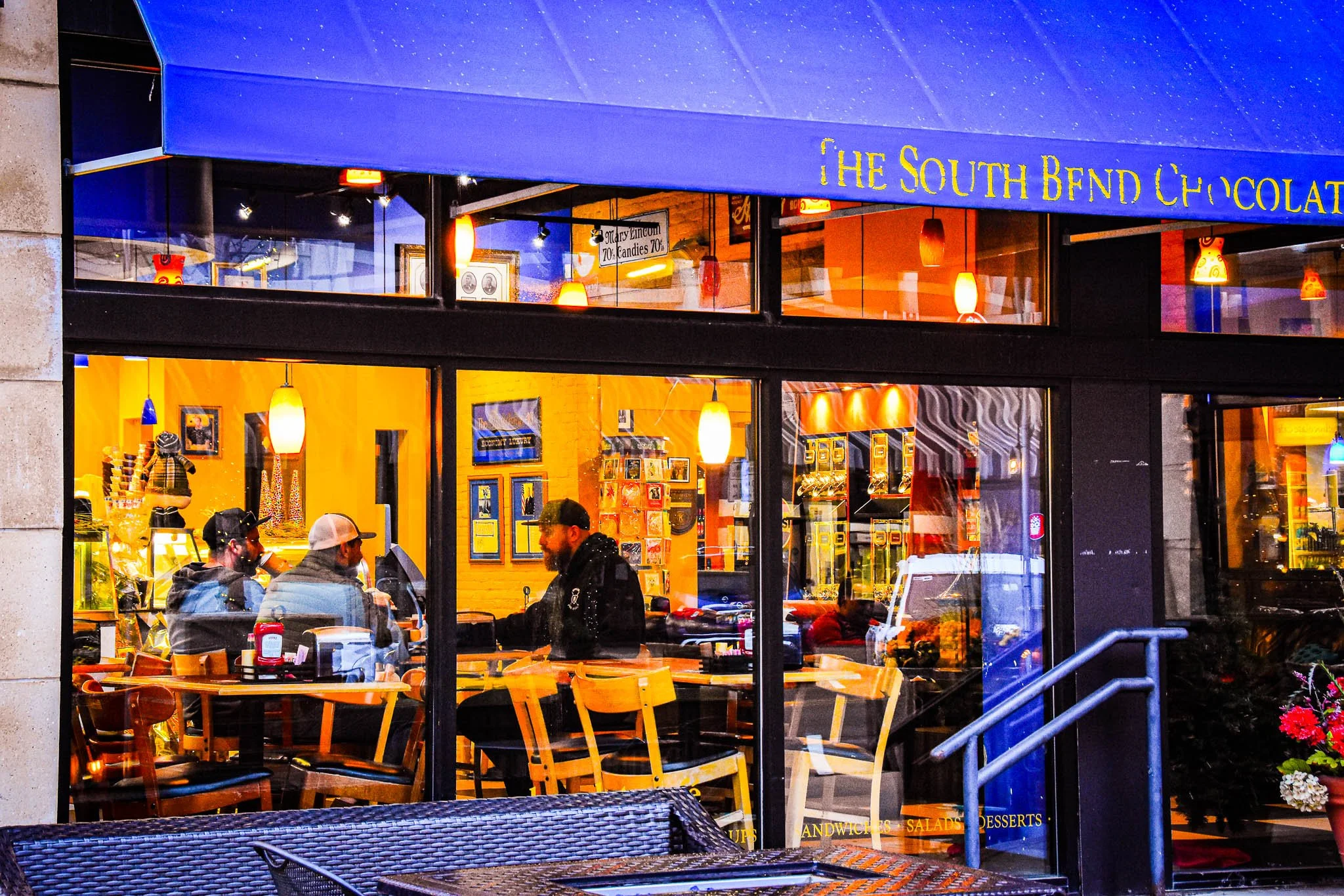 Inside a cozy cafe with yellow walls and warm lighting, three people are sitting at a table engaged in conversation, with a barista behind the counter. The exterior has large glass windows, and a blue awning with gold lettering reads 'The South Bend 