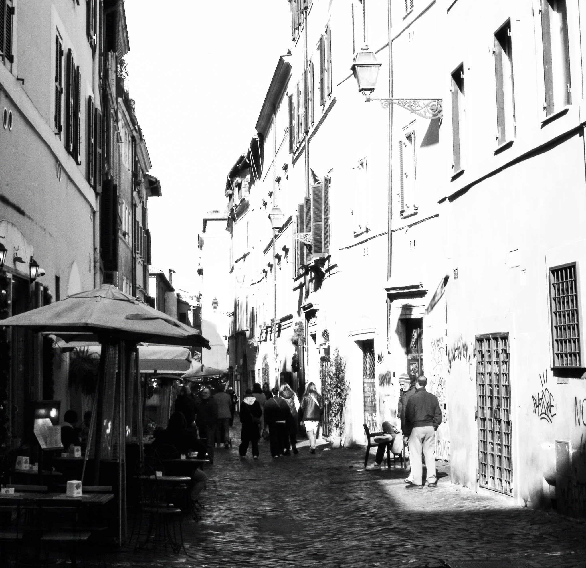 A black-and-white photo of a narrow European city street with outdoor seating, pedestrians walking, and old buildings with shuttered windows.