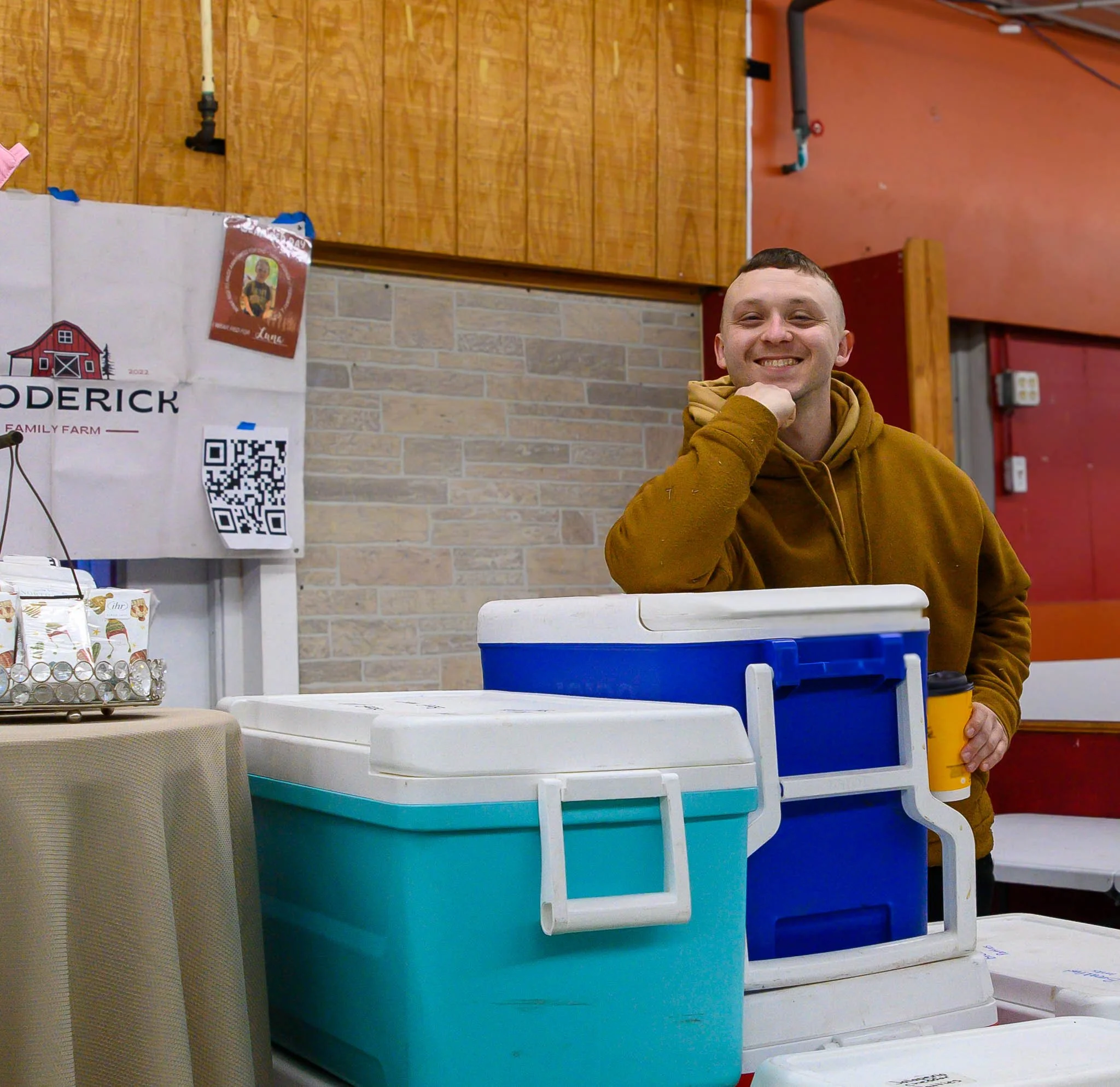 A smiling man in a brown hoodie standing behind two coolers, one blue and one turquoise, at a booth. He is holding a yellow cup and resting his chin on his hand. The background features a brick wall, wooden paneling, and a banner for Derrick Family F