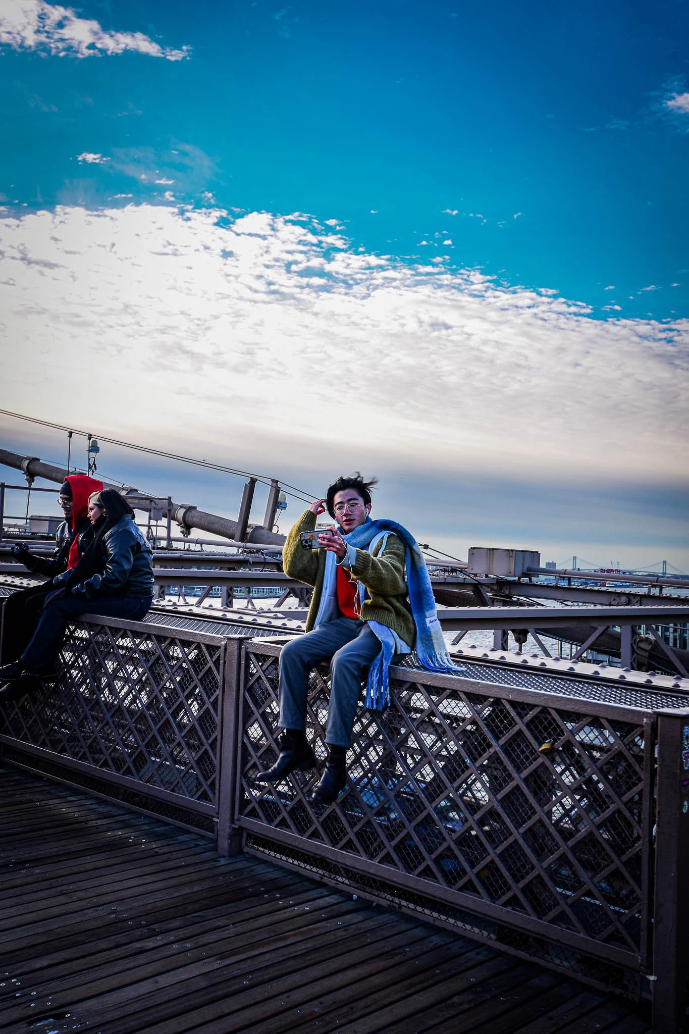 Three people sitting on the railing of a bridge with a cloudy sky in the background. One woman is holding a phone and looking at the camera, wearing a colorful scarf, green jacket, and glasses. The other two individuals are sitting nearby, one wearin