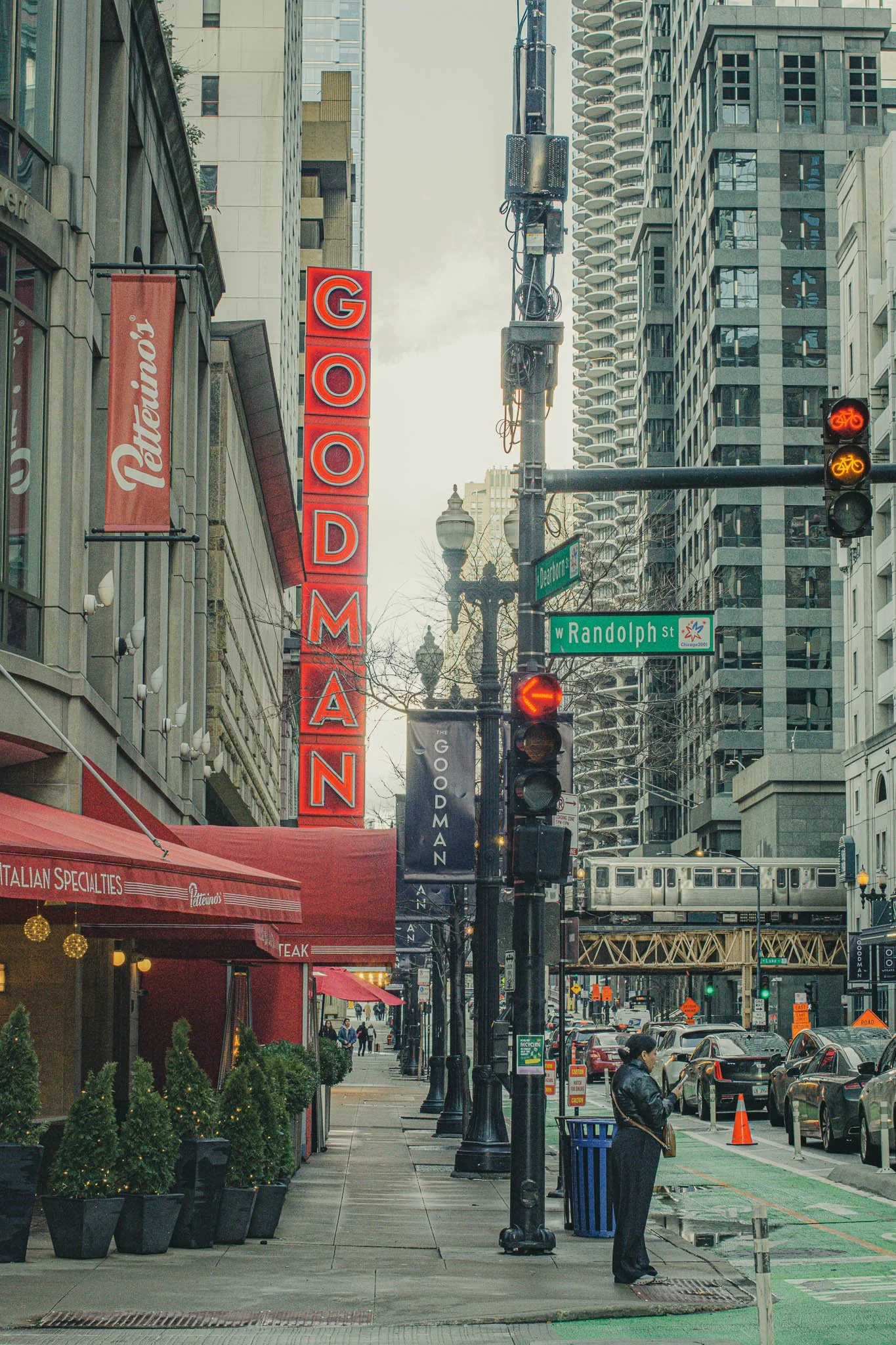 City street scene with a sign reading 'GODFATHER' and building signs for 'Peterson', street signs for 'Randolph St' and 'Dearborn St', a train crossing above the street, and a woman standing at a street corner looking at her phone.