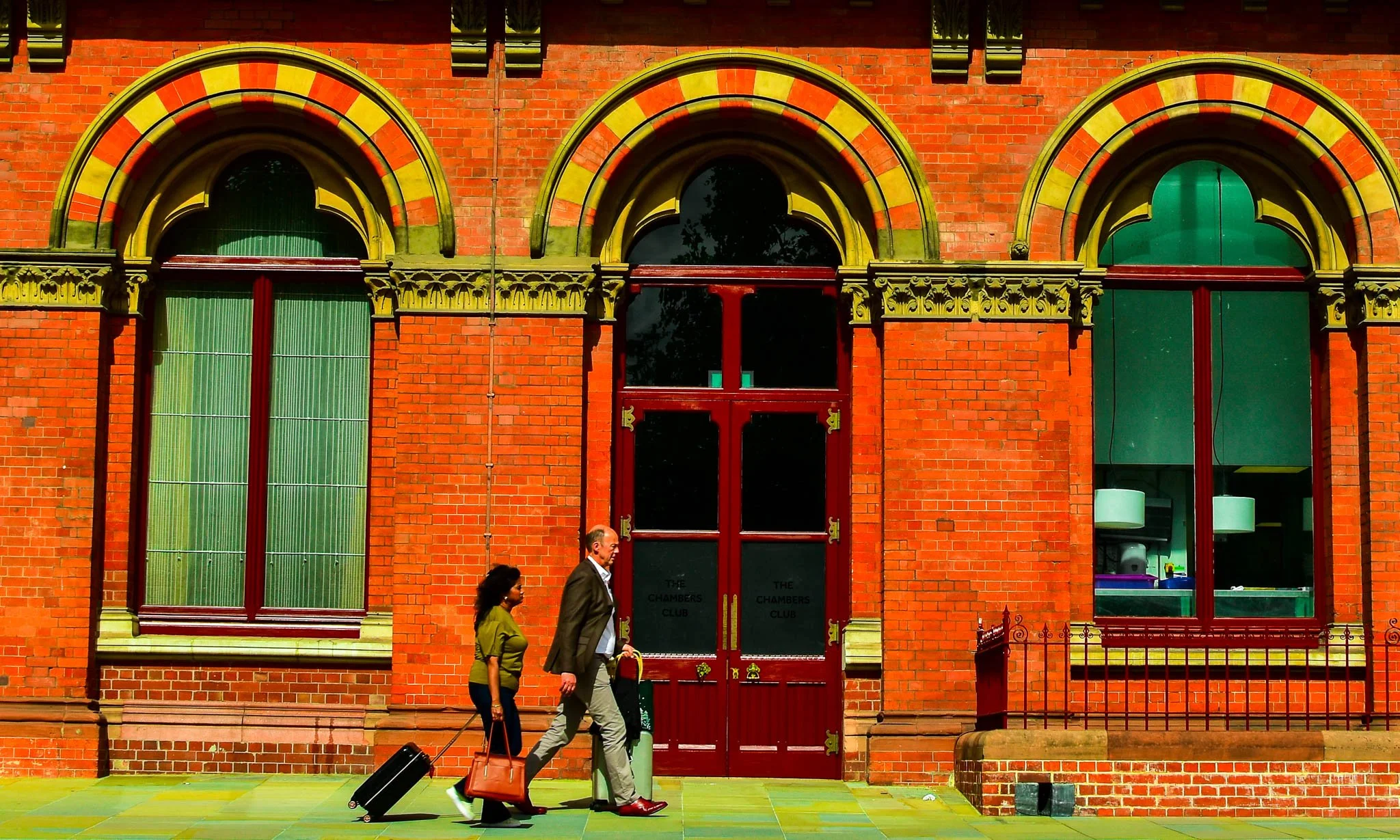 Two people walking past a red brick building with tall arched windows and double doors. One person pulls a small rolling suitcase, and the other carries a brown bag.