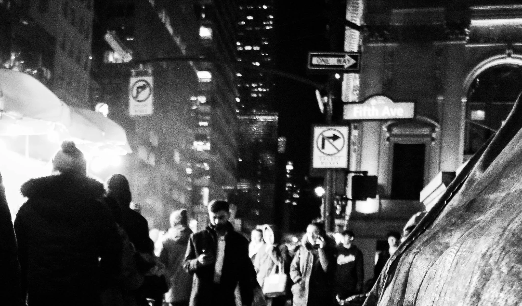 Black and white photo of a busy city street at night with people walking and street signs, including one for Fifth Avenue.