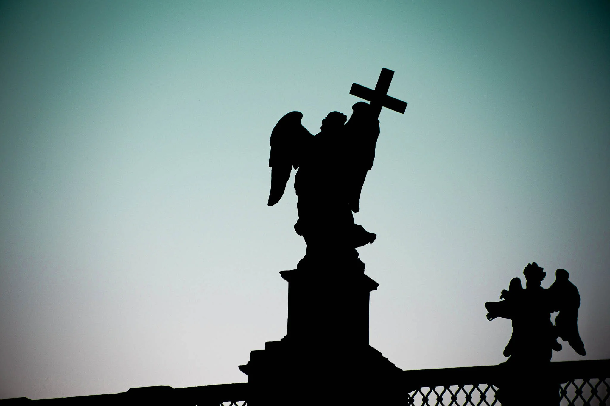 Silhouette of a religious statue holding a cross, with smaller figures nearby, against a light sky.