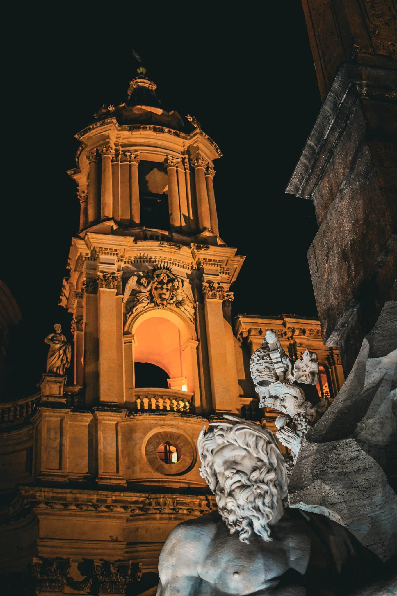Nighttime photo of a historic building with ornate architecture, illuminated with warm lighting, and a sculpture of a bearded man in the foreground.