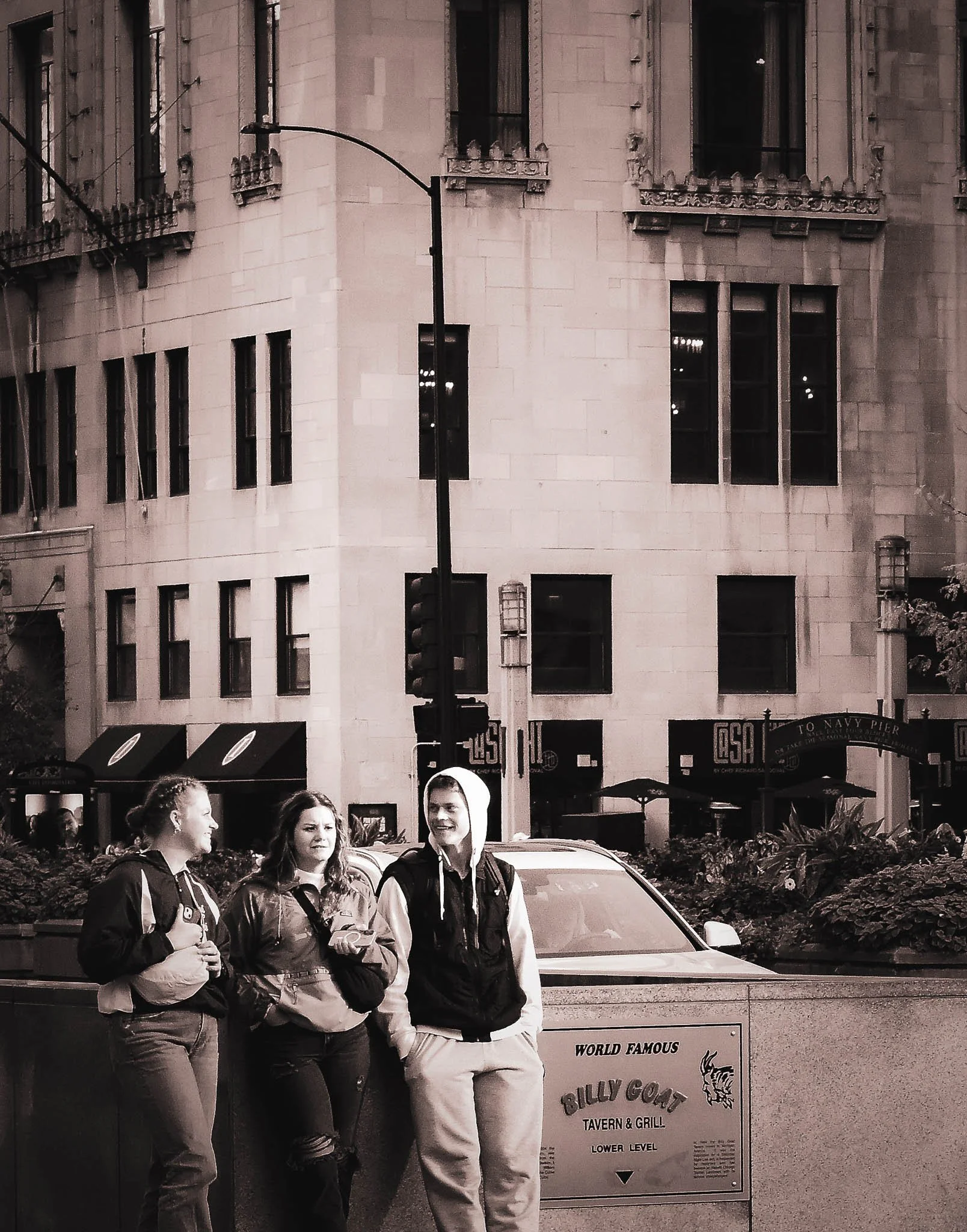 Three young people stand together on a city sidewalk near a sign for Billy Goat Tavern & Grill, with a tall building and street in the background.