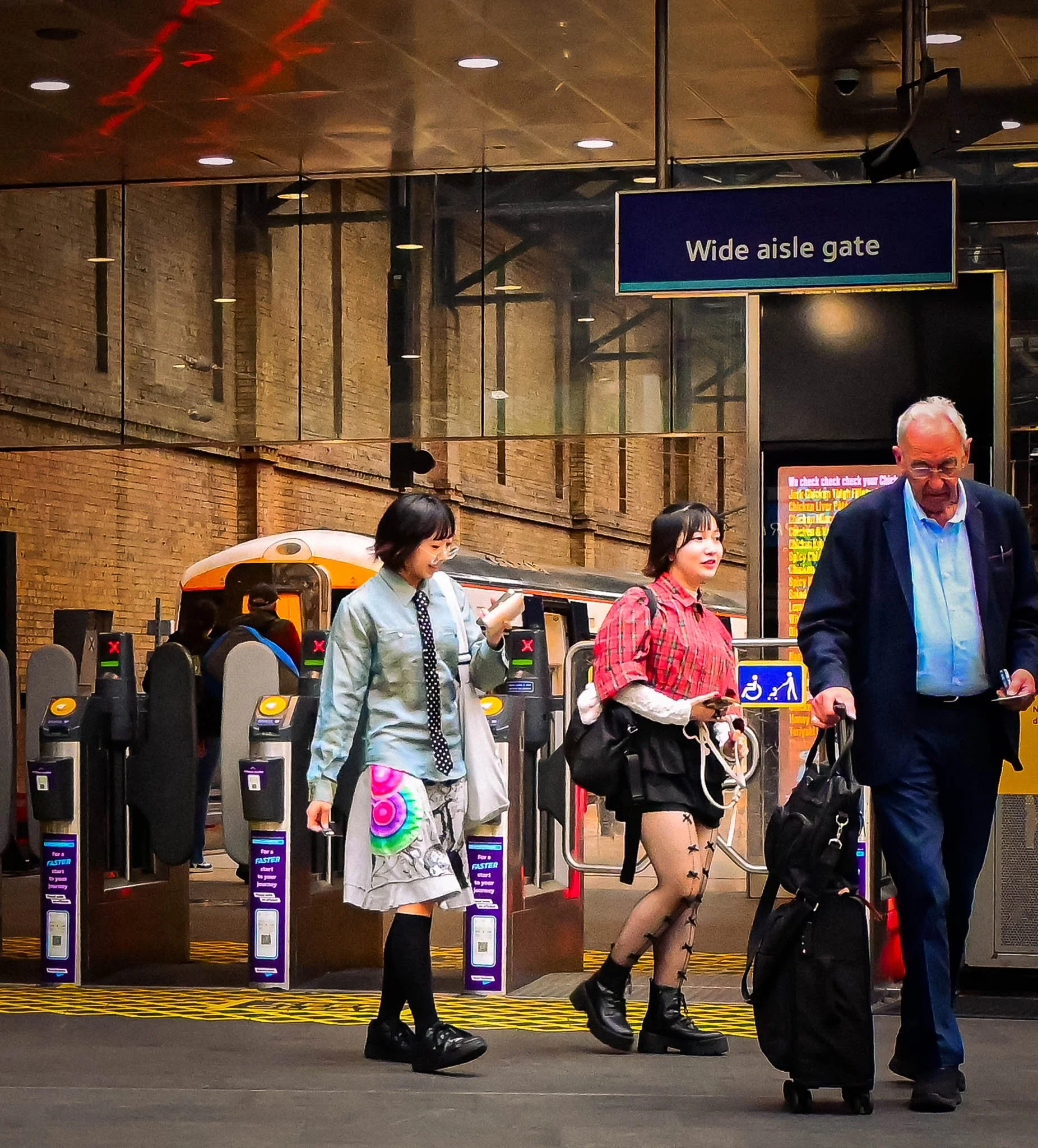 People walking through a train station near wide aisle gate, some with luggage and backpacks, with a train in the background and signs overhead.
