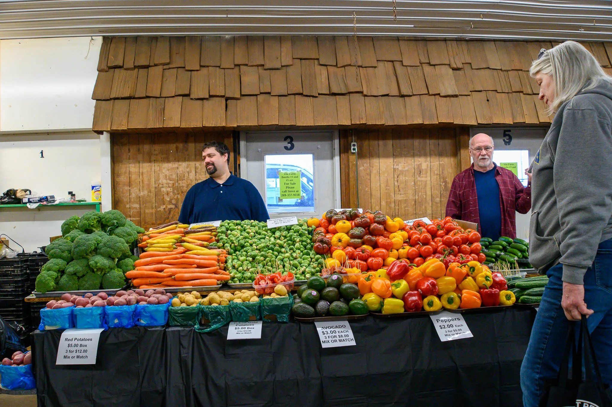 A farmer's market stall with various fresh vegetables, including broccoli, carrots, Brussels sprouts, potatoes, and an assortment of colorful peppers. Two male vendors stand behind the stall, and a female customer is in front, looking at the produce.