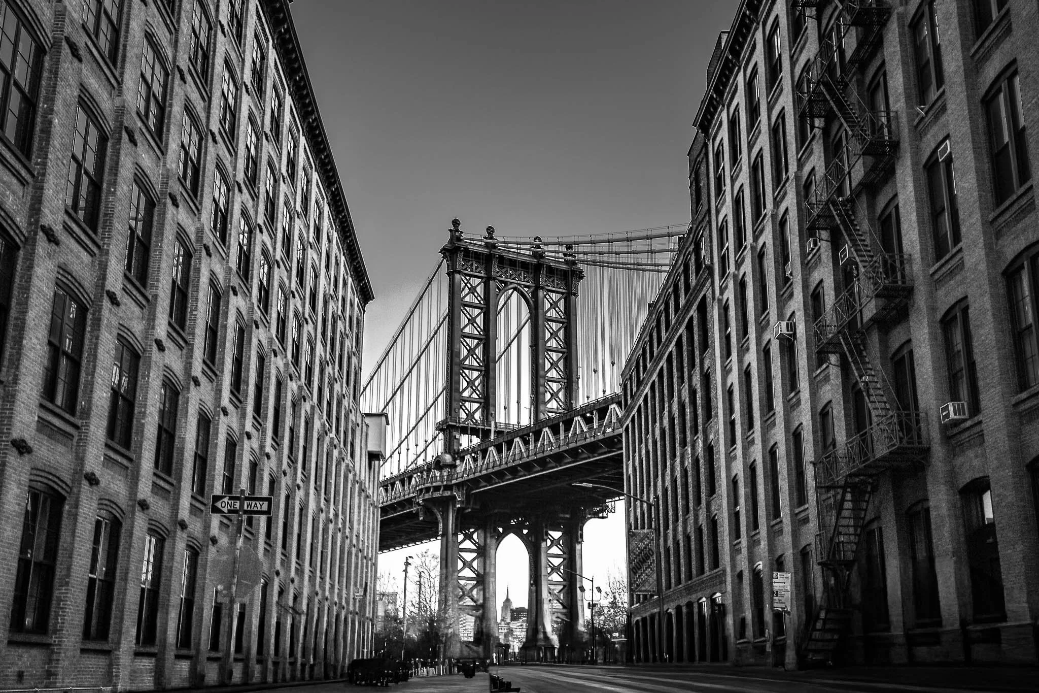Black and white photo of Manhattan Bridge viewed from a narrow street flanked by tall brick buildings with fire escapes.