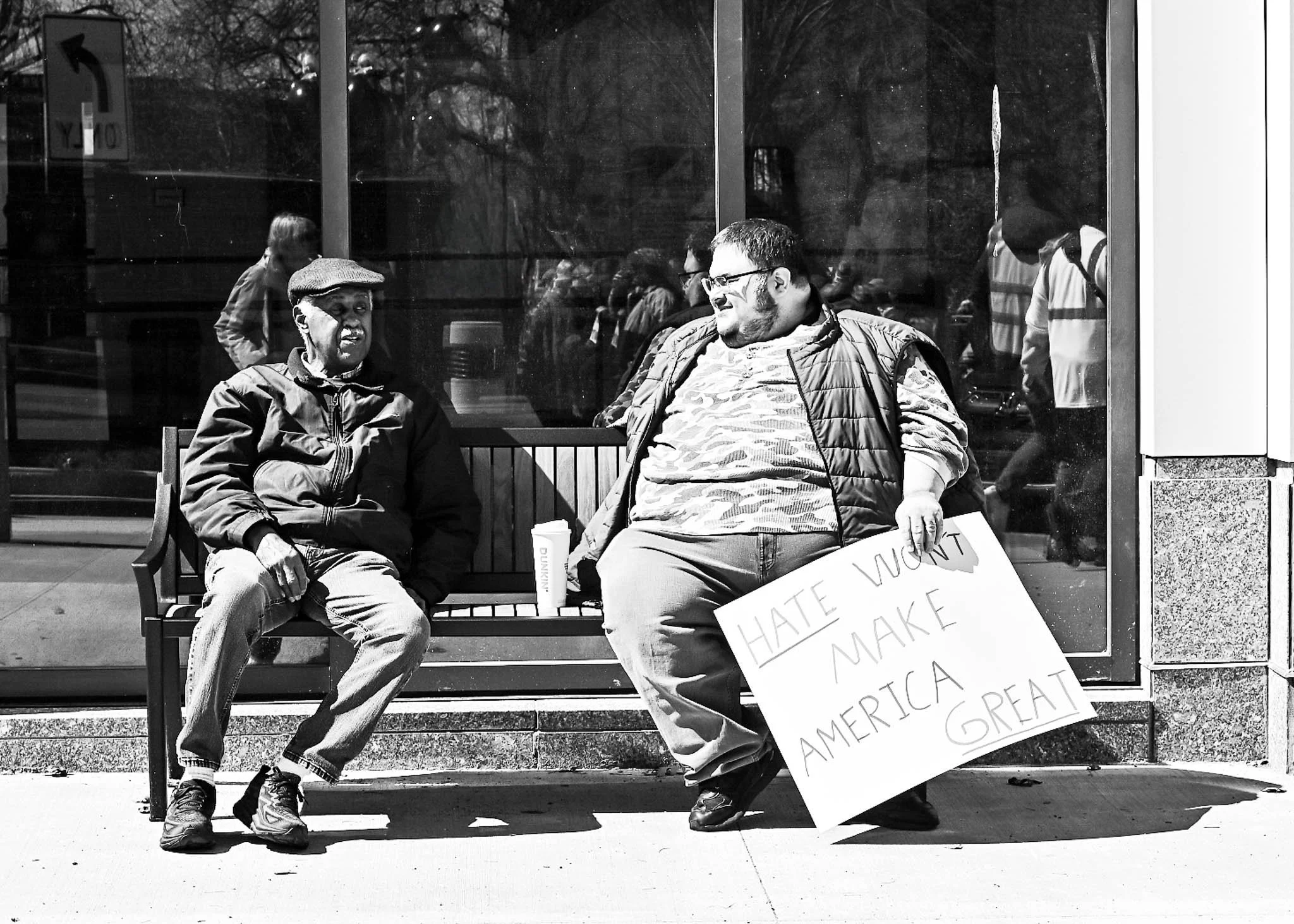 Two men sit on a bench outside a building, one holding a sign that reads 'Hate won't make America great'. The man on the left wears a cap and jacket, while the man on the right wears glasses, a vest, and a camo shirt.