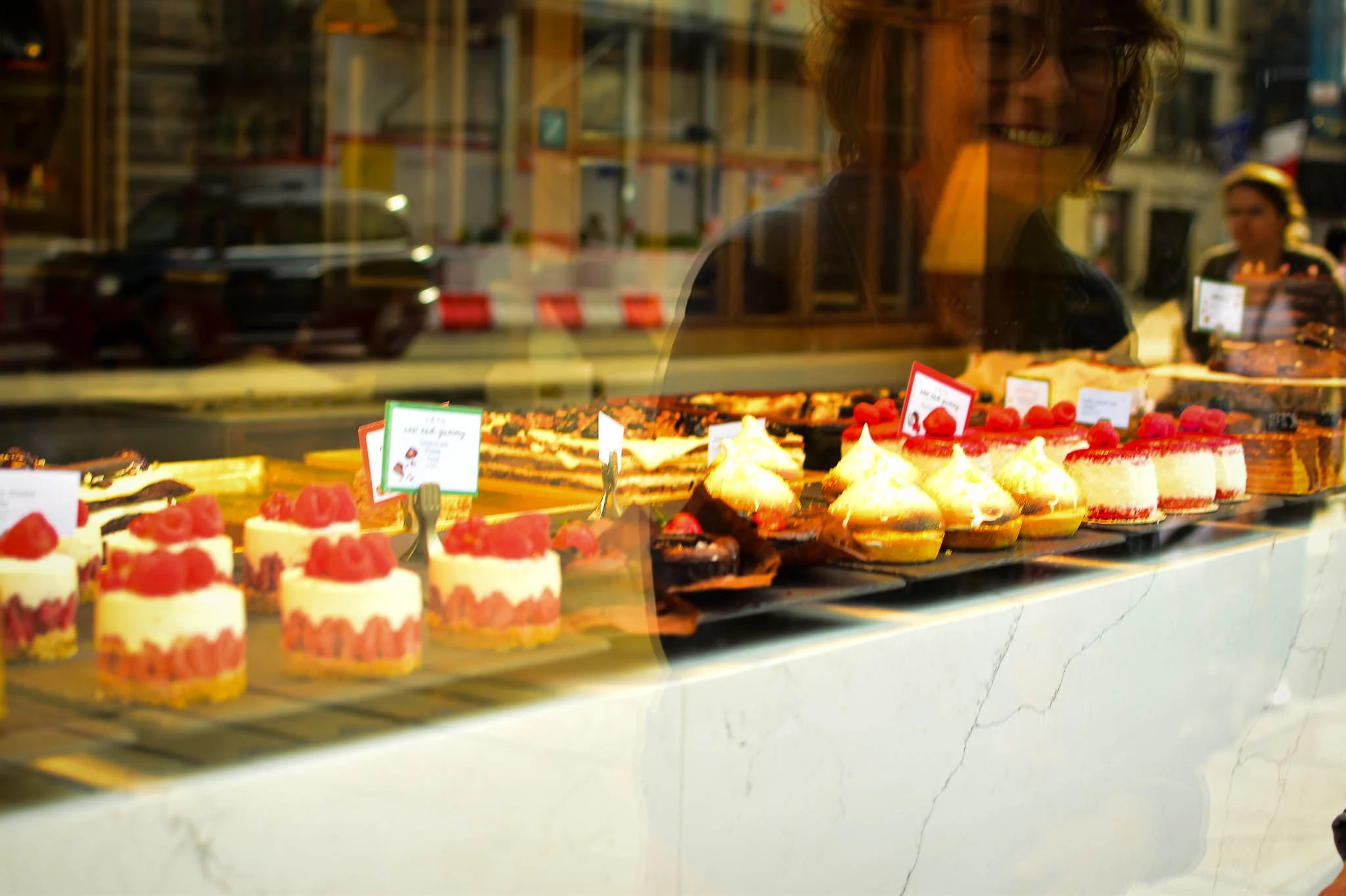 A display of various cakes and desserts in a bakery window, with a smiling person behind the glass and a reflection of the street outside.