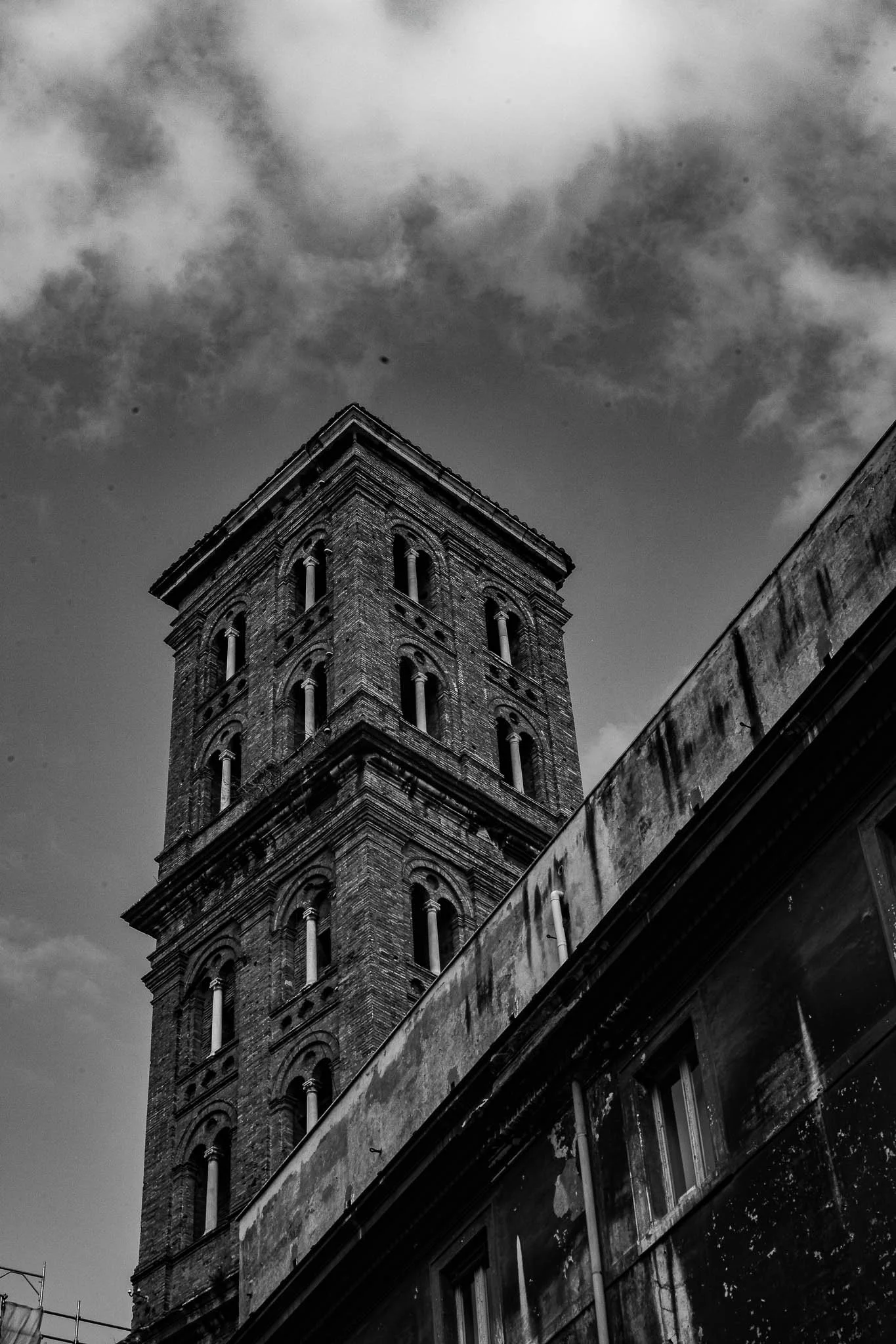Black and white photo of an old brick tower with arched windows, part of an urban building, with a cloudy sky in the background.