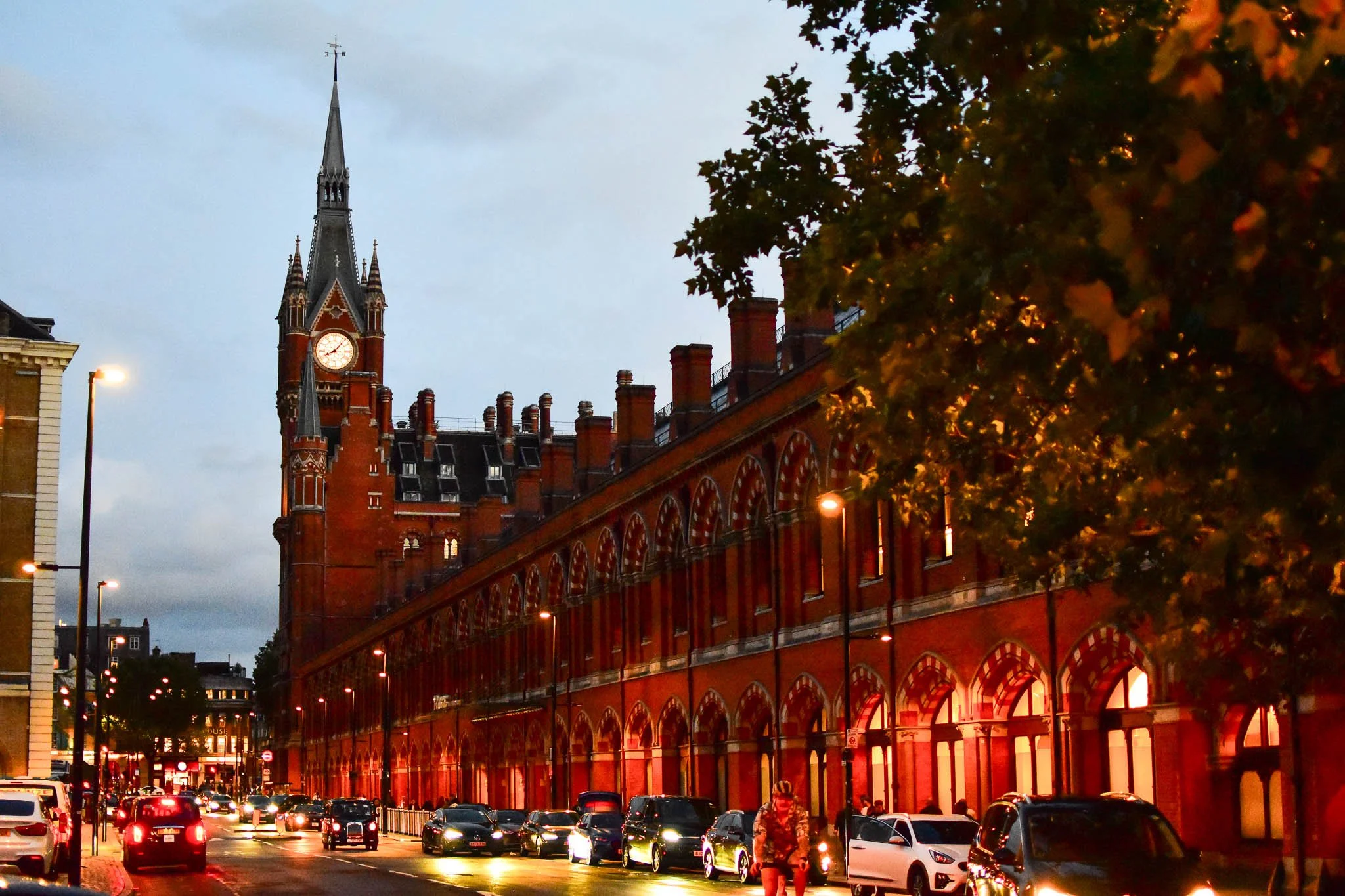 Street view of a red brick building with a clock tower at dusk, streetlights, cars, and pedestrians.