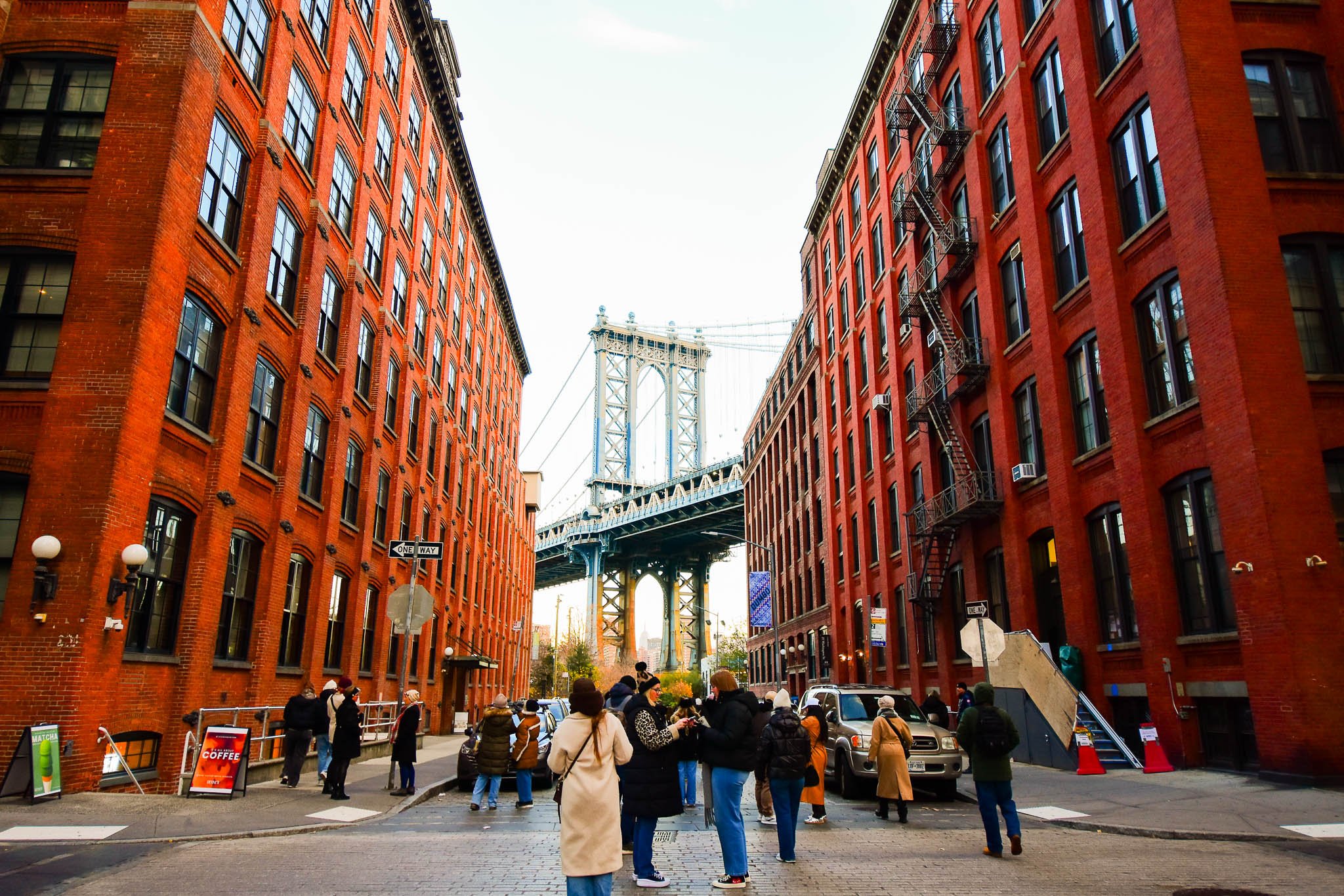 People walking on a city street with red brick buildings on both sides and a suspension bridge in the background.