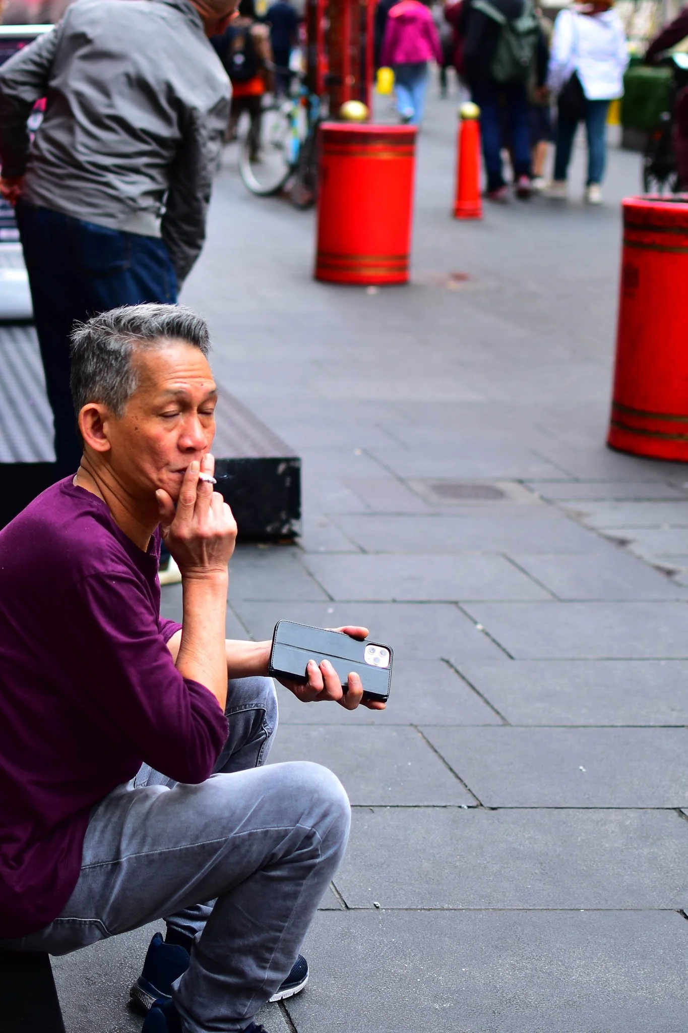 A man sits on a step outside on a busy city street, smoking a cigarette and holding a phone, with people walking in the background.