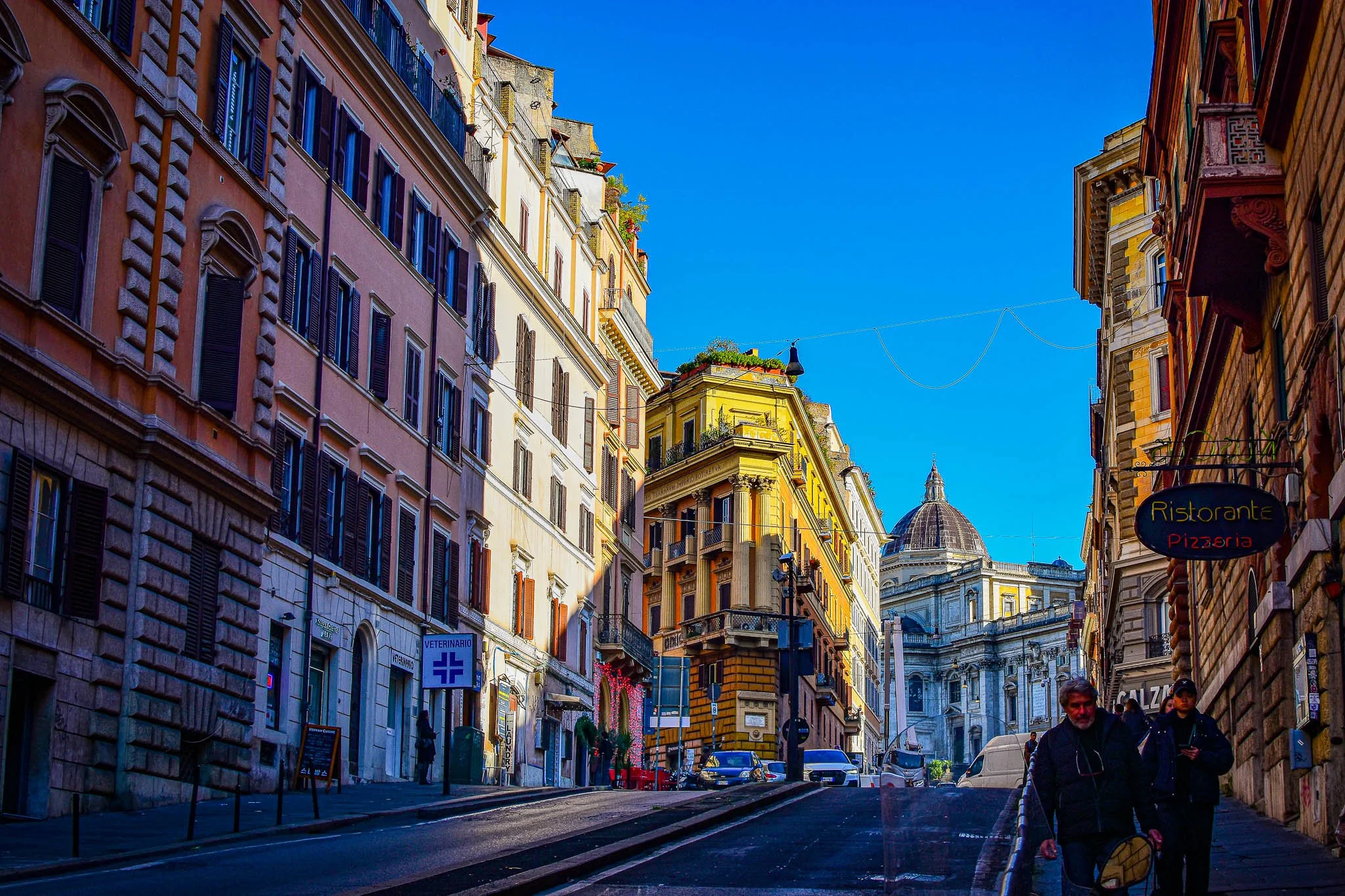 A city street in Rome with colorful buildings, a church in the background, and pedestrians walking.