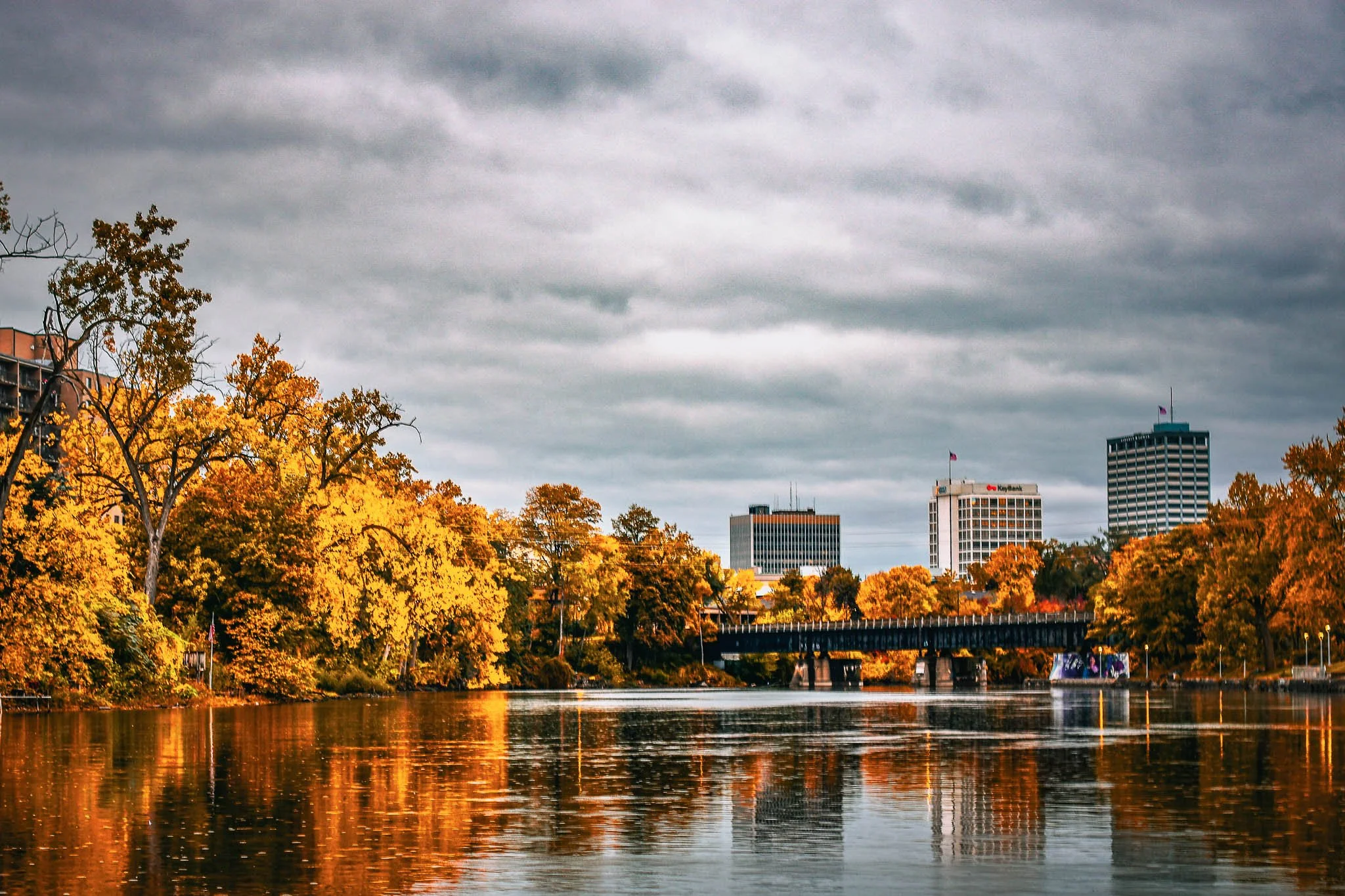 City skyline with tall buildings and trees with orange and yellow leaves reflecting in the river, under a cloudy sky.