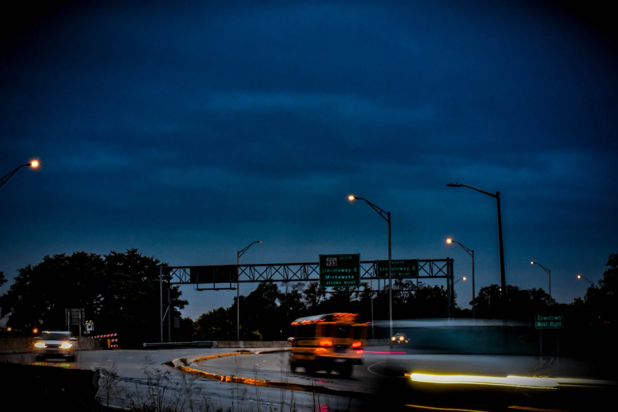 Nighttime highway scene with moving vehicles and illuminated streetlights, overhead freeway signs for Lincolnway E and Mishawaka, Southbound, near downtown.