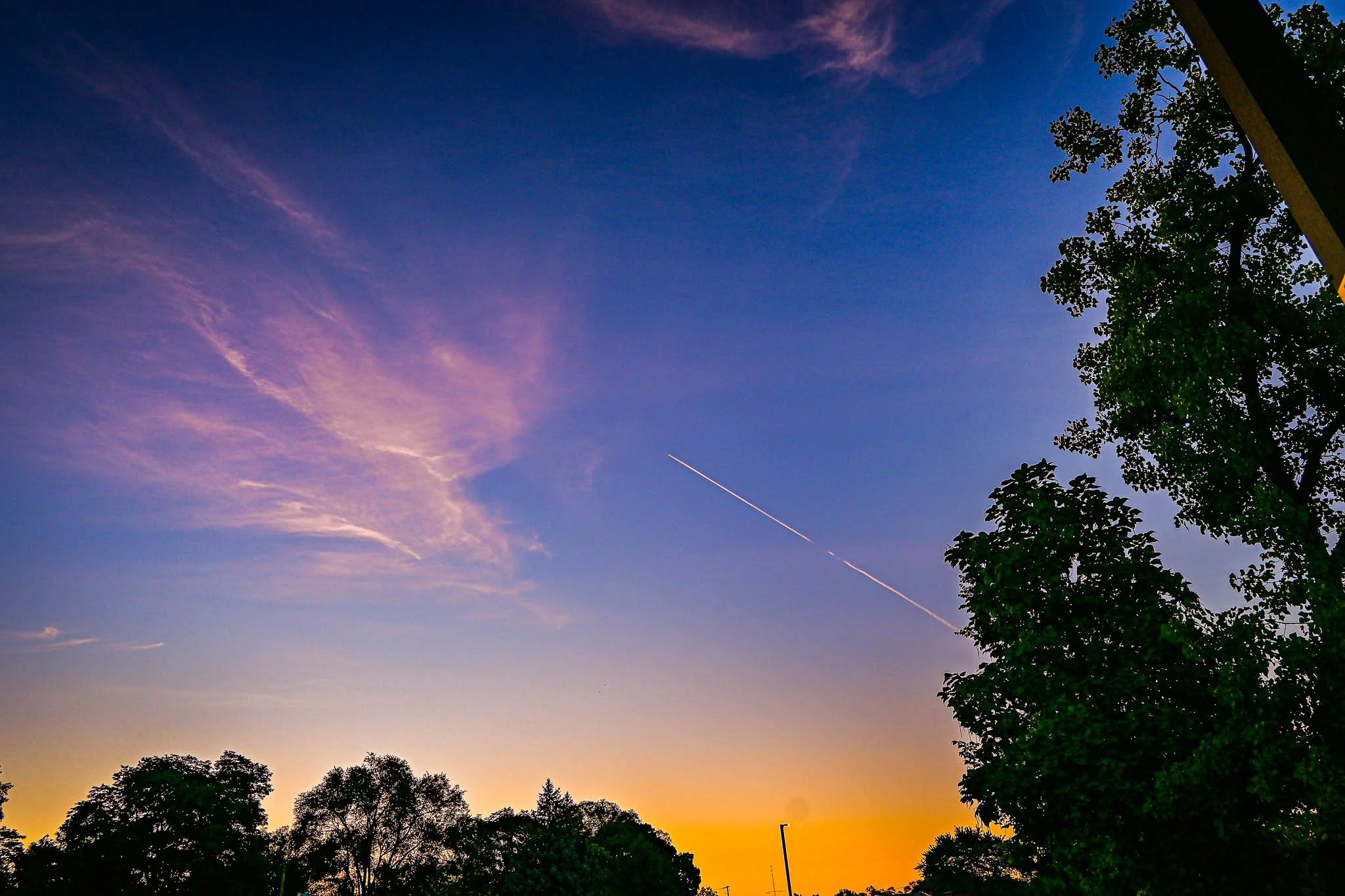 A colorful sunset sky with orange near the horizon and transitioning to purple and blue higher up, partly cloudy with wispy pink clouds, trees silhouetted in the foreground, and a faint contrail from an airplane crossing the sky.