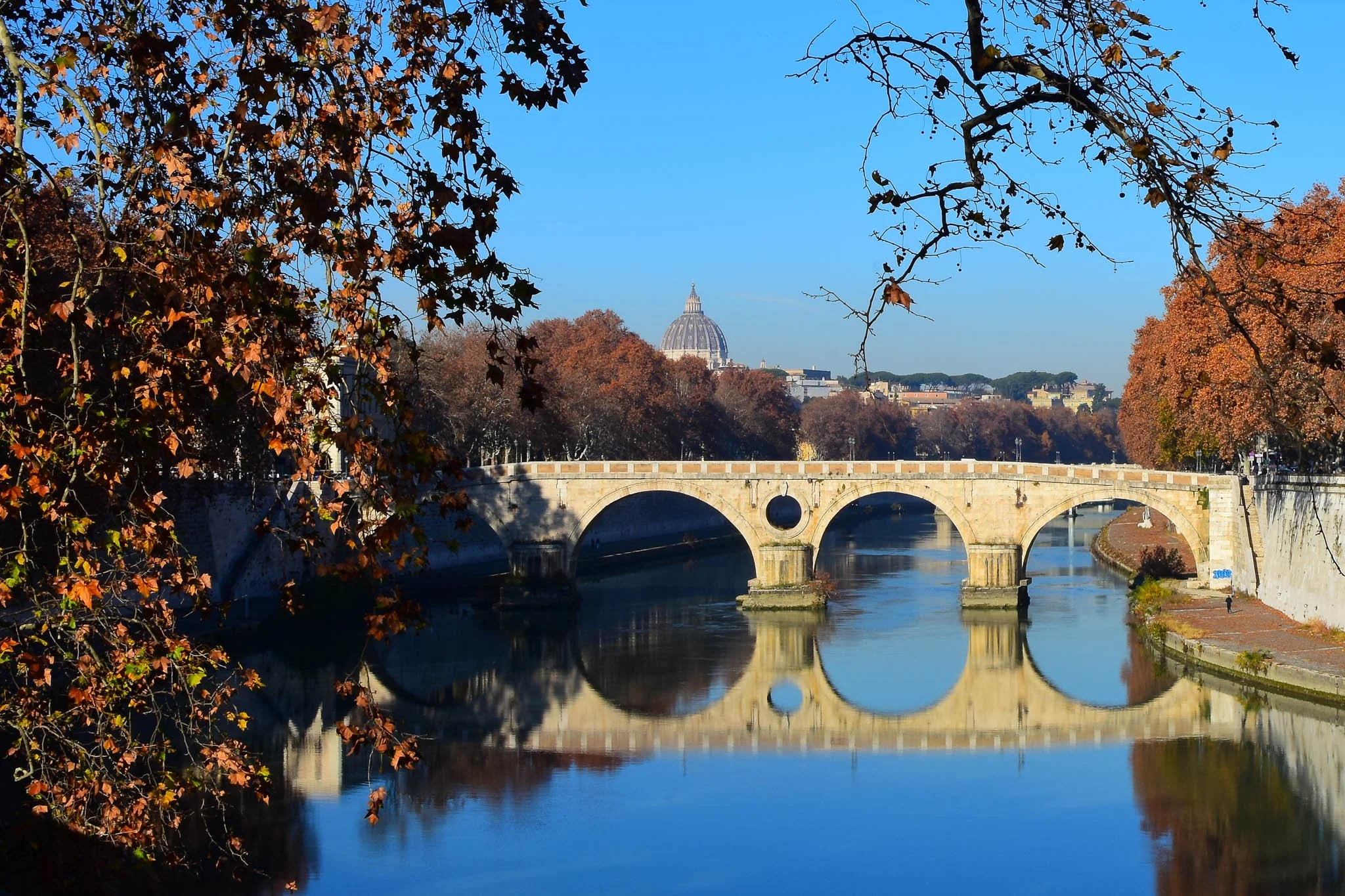 A scenic view of a stone bridge over a calm river with reflections, framed by autumn-colored trees, and a domed building in the background on a clear blue sky day.