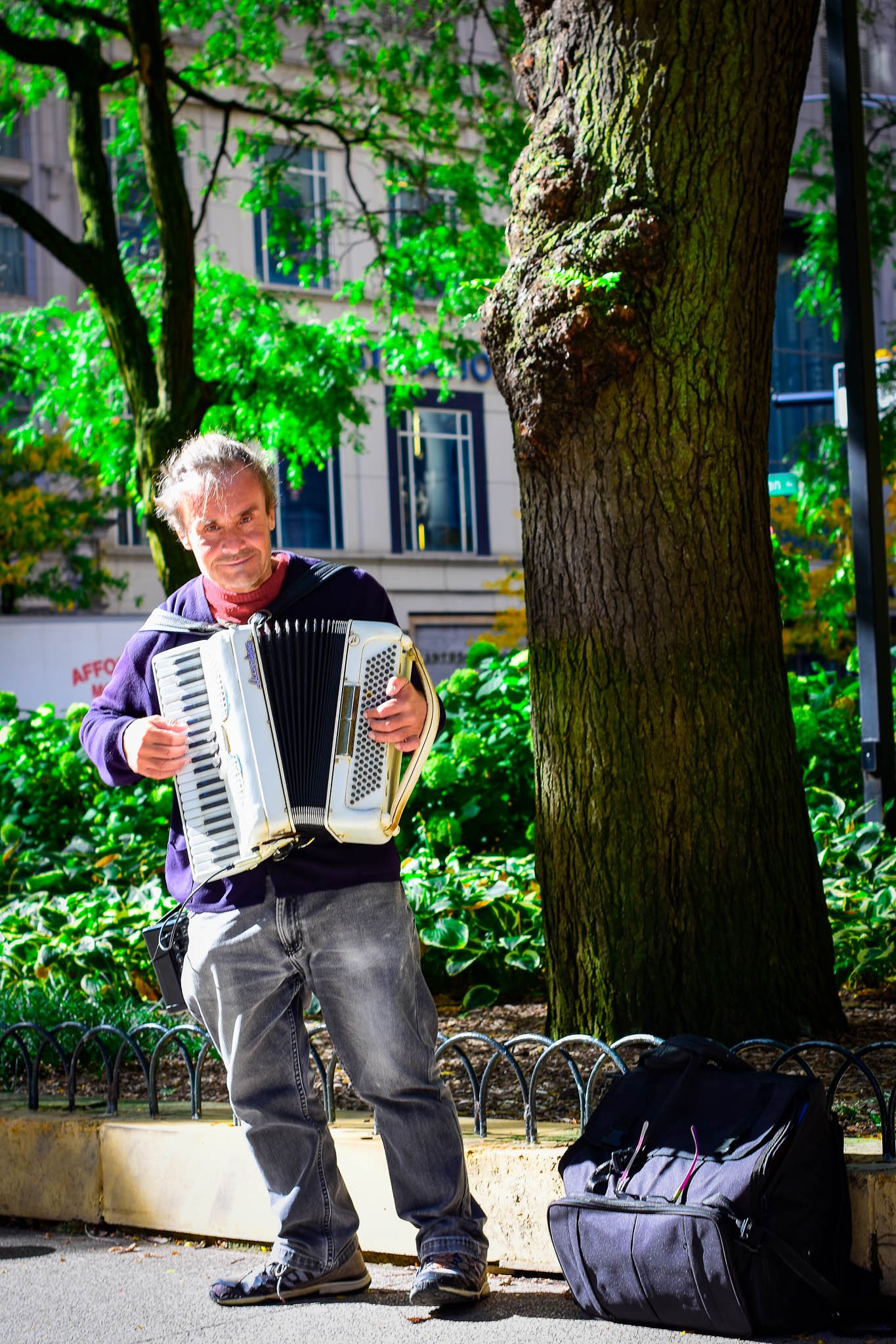 A man playing an accordion on a city sidewalk with trees and buildings in the background, and a black backpack on the ground beside him.