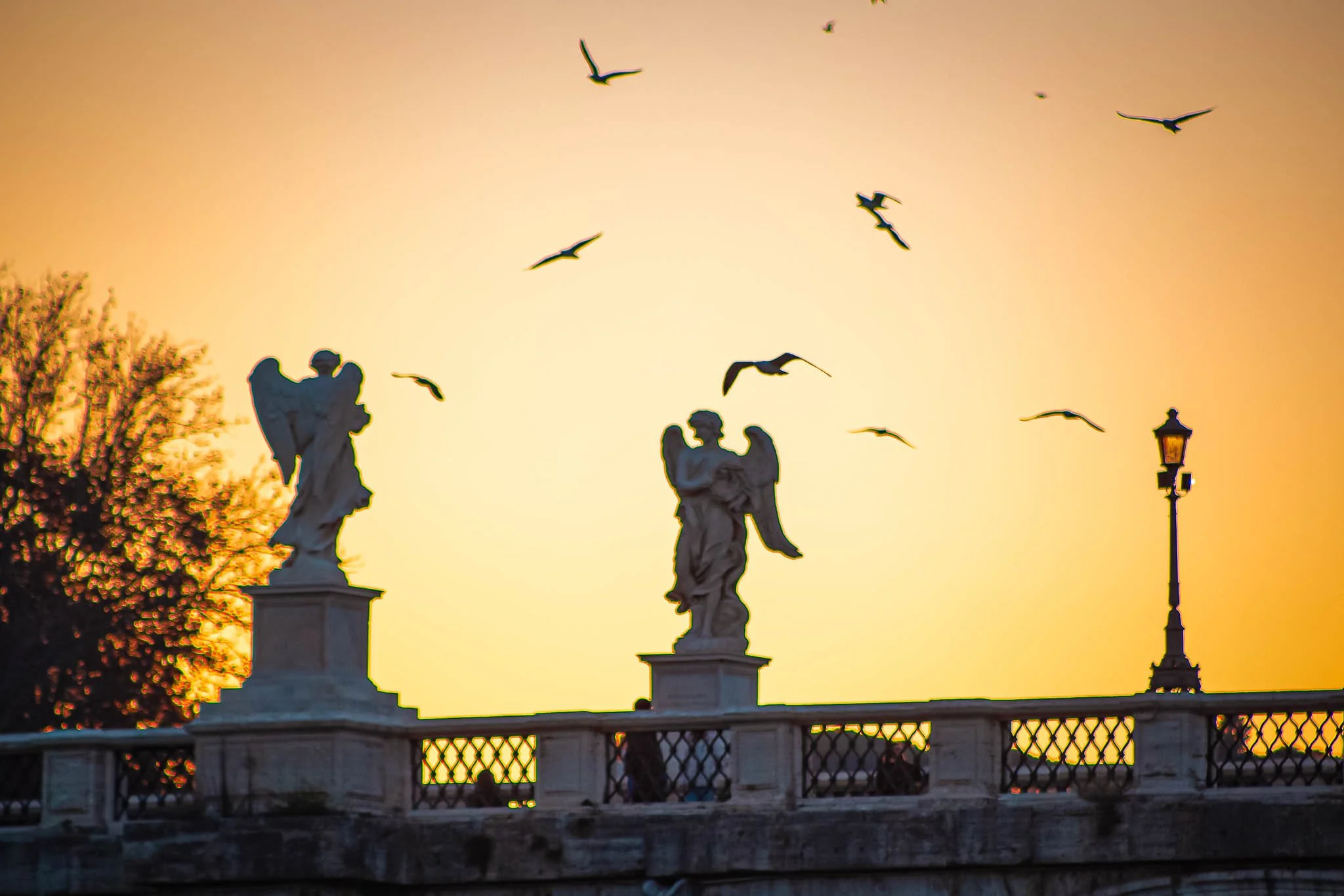 Silhouetted statues of angels on a bridge during sunset with birds flying overhead and a street lamp on the right.