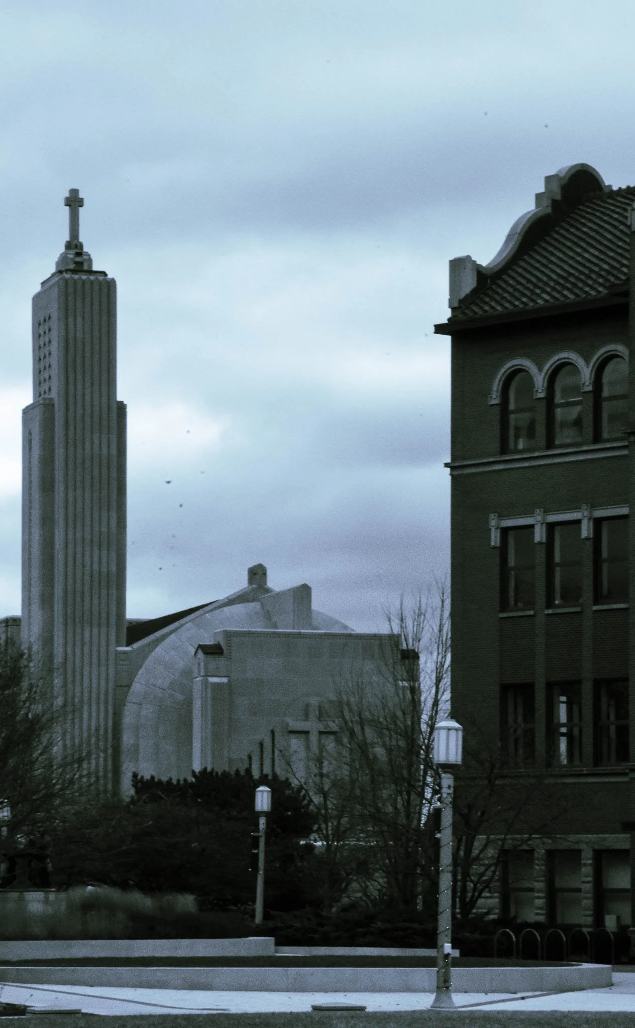 Cityscape featuring a tall building with a cross on top, a church or religious structure, and older brick buildings, under a cloudy sky.
