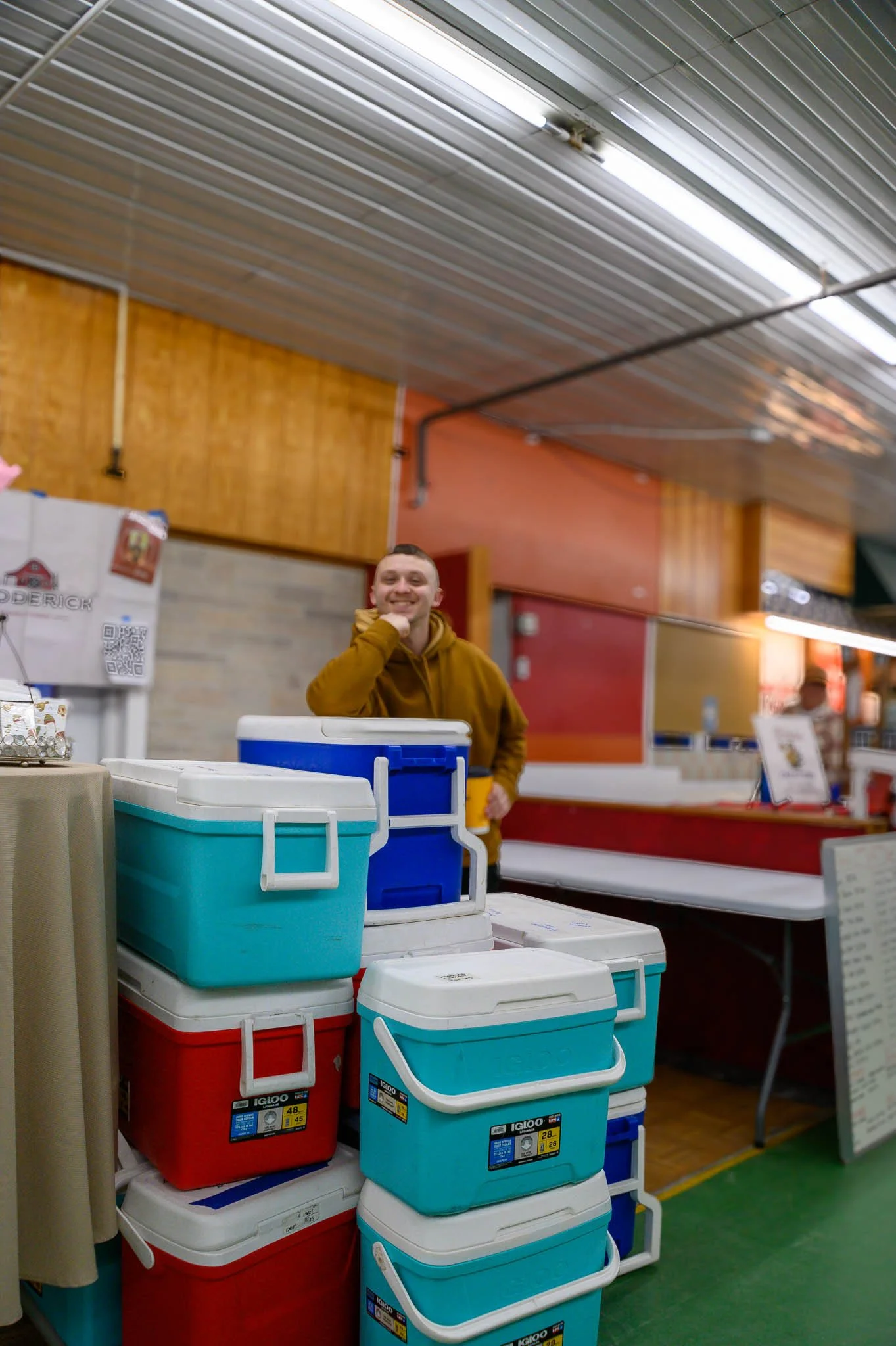 A man smiling behind a stack of colorful coolers inside a store with a wooden ceiling and walls.