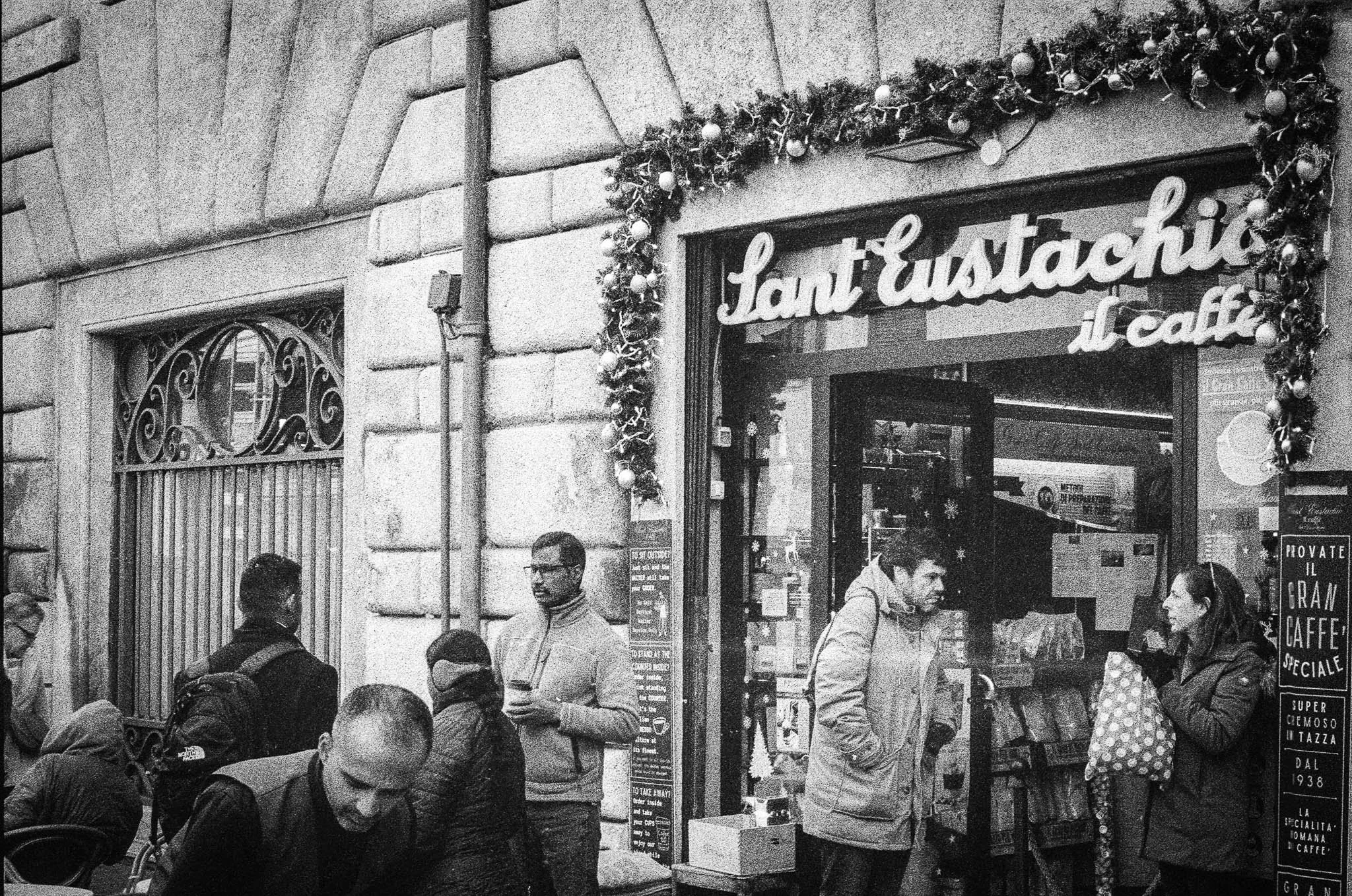 People standing outside Saint Eustachio il Caffè, decorated with Christmas garland, on a city street.
